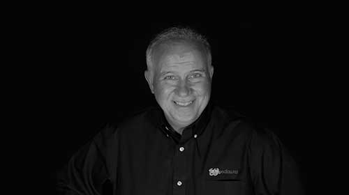 A person smiling in a black-and-white portrait, wearing a buttoned shirt with a logo, set against a dark background.