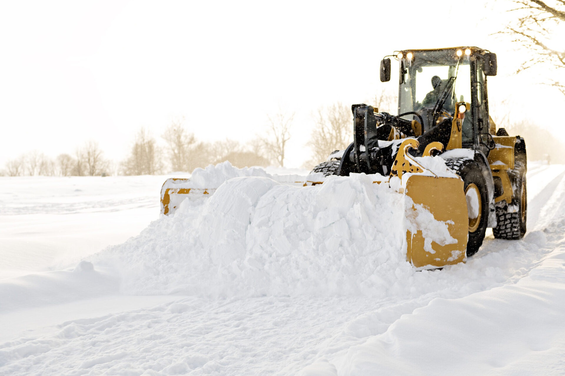A person operates a snowplow clearing heavy snow on a road, surrounded by a snowy landscape and barren trees on a bright day.