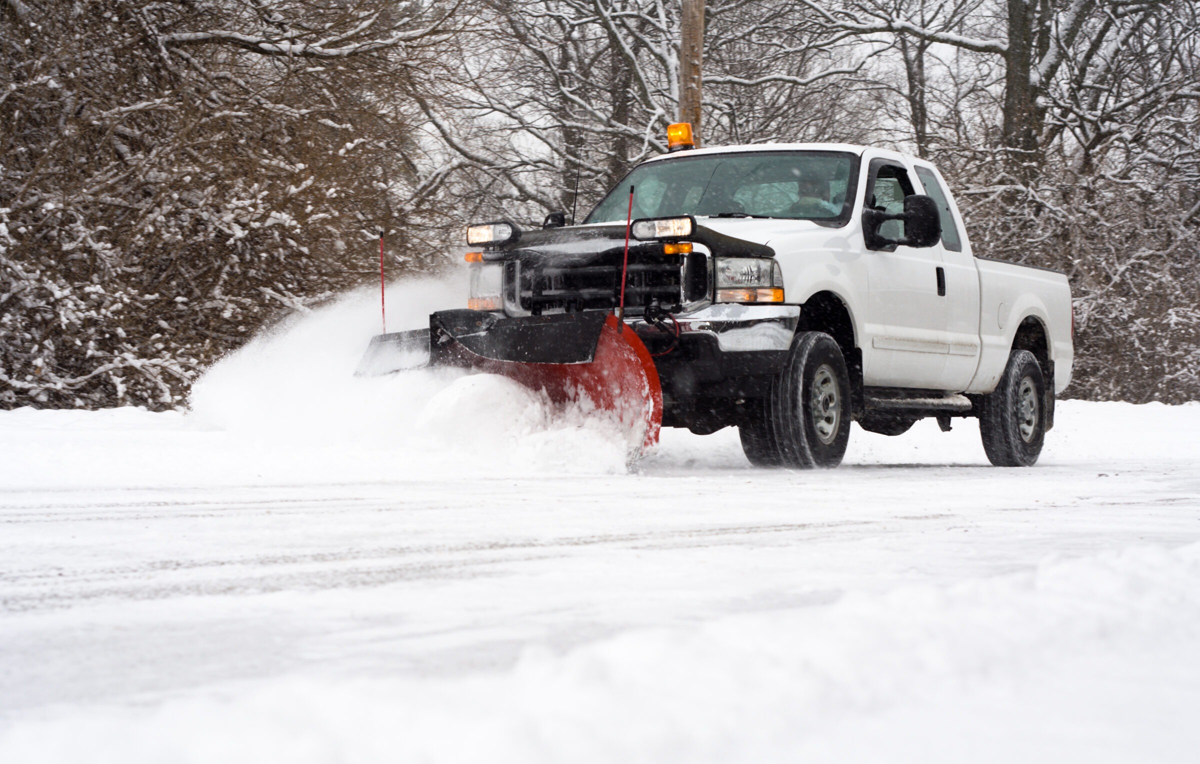 A white truck with a snowplow clears a snowy road lined with bare trees, creating a path through the winter landscape.