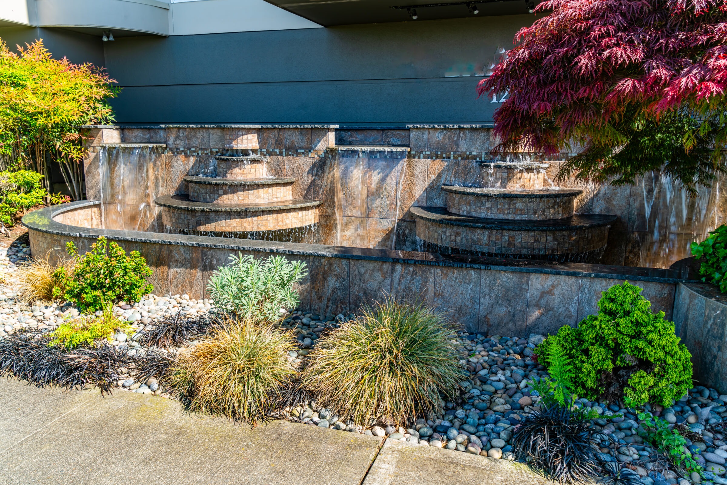 A layered, stone fountain features cascading water, surrounded by lush green plants and colorful foliage, set against a modern building facade.