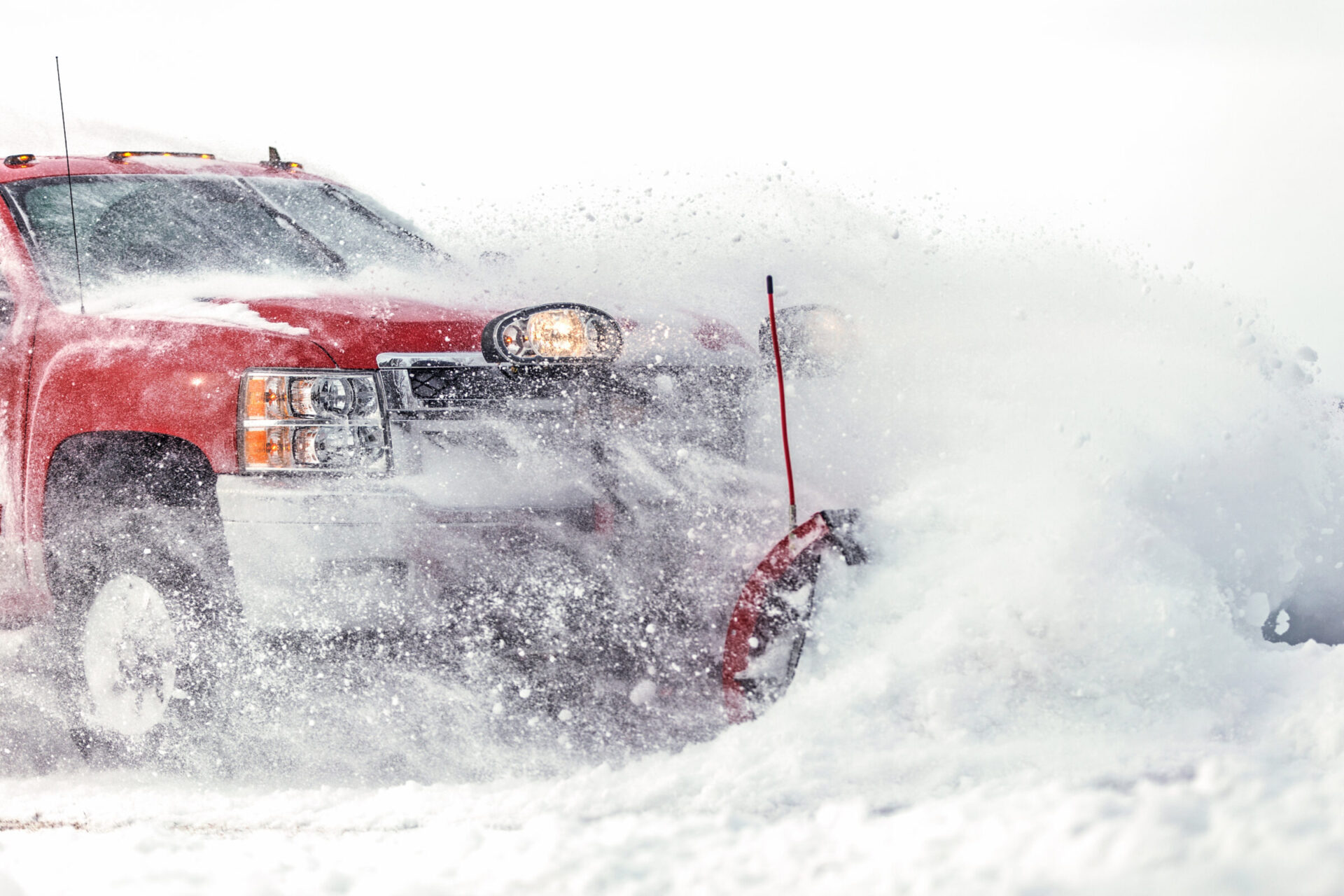 A red truck plowing through snow, clearing a path. Bright headlights illuminate swirling snow, creating a dynamic, wintery scene.