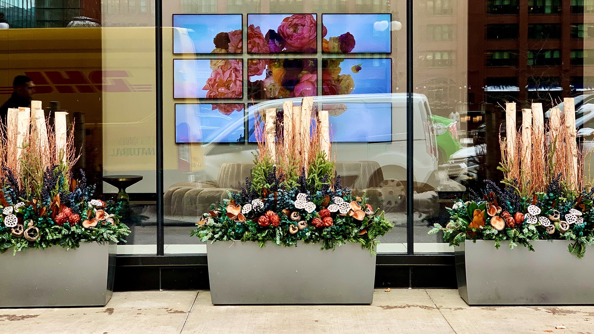 Three large planters with vibrant floral arrangements in front of a glass window, reflecting urban street scene with passing vehicles.