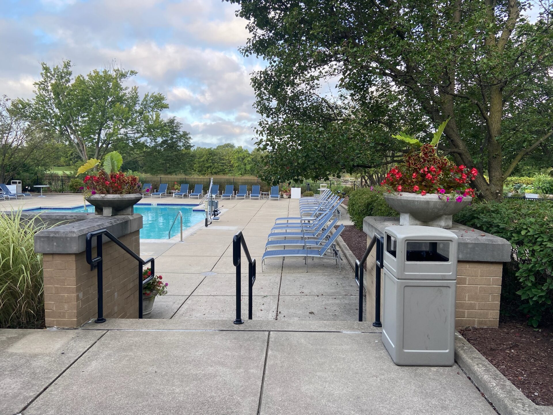 A serene outdoor pool area with lounge chairs, surrounded by trees and flowers, under a partly cloudy sky.