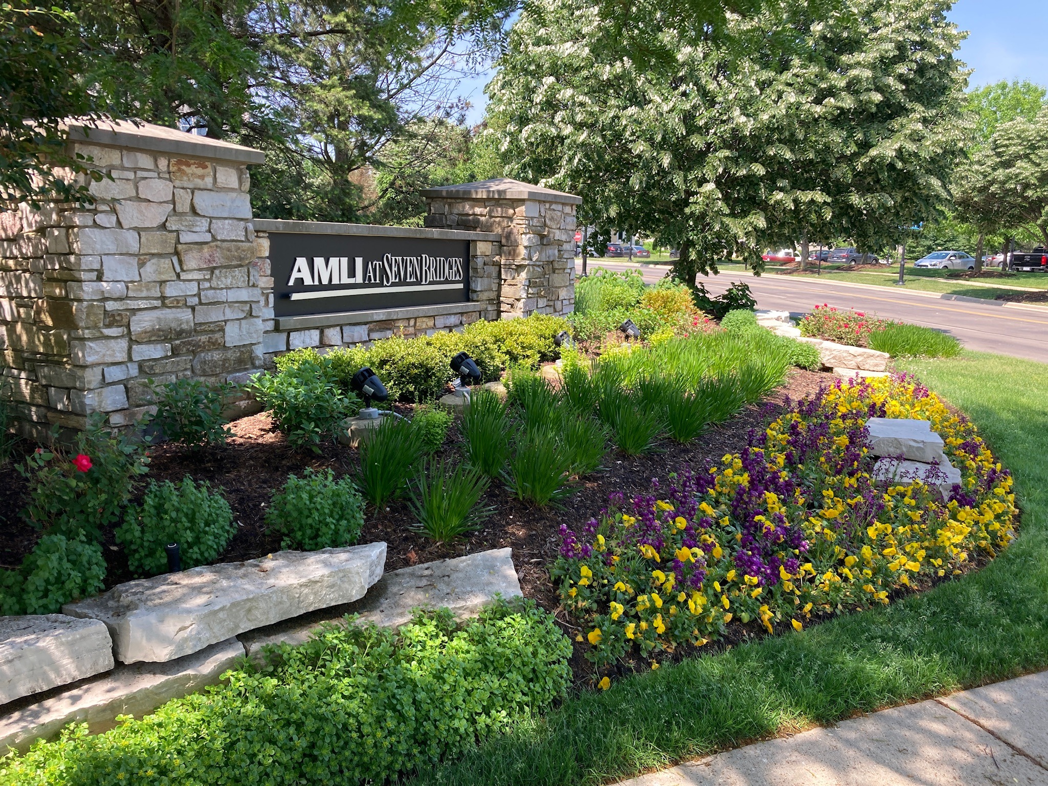Stone sign for "AMLI at Seven Bridges" surrounded by colorful flowers, greenery, and trees; a road is visible in the background.