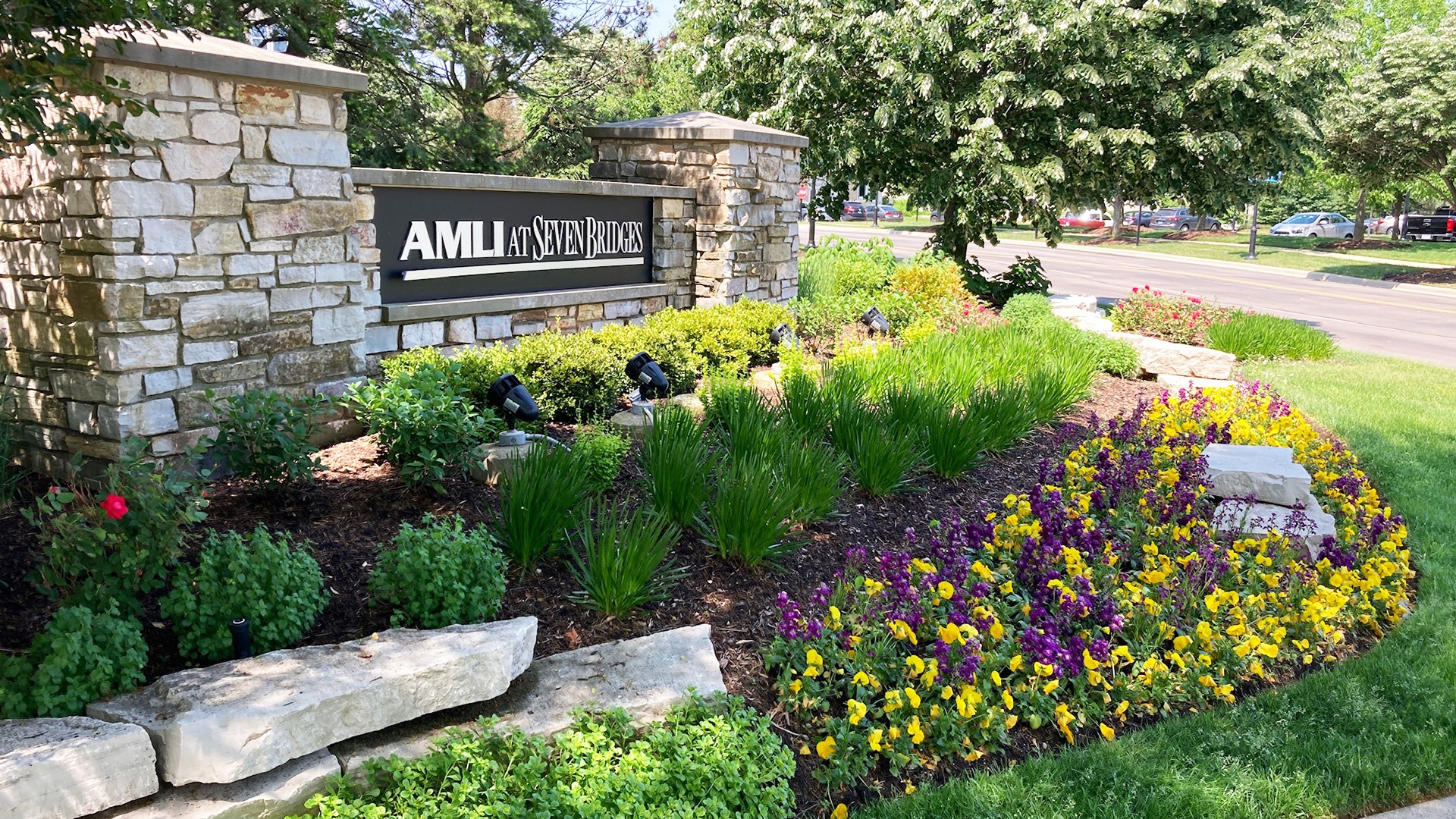 Stone sign reads "AMLI at Seven Bridges," surrounded by vibrant flowers and greenery along a road in a park-like setting.