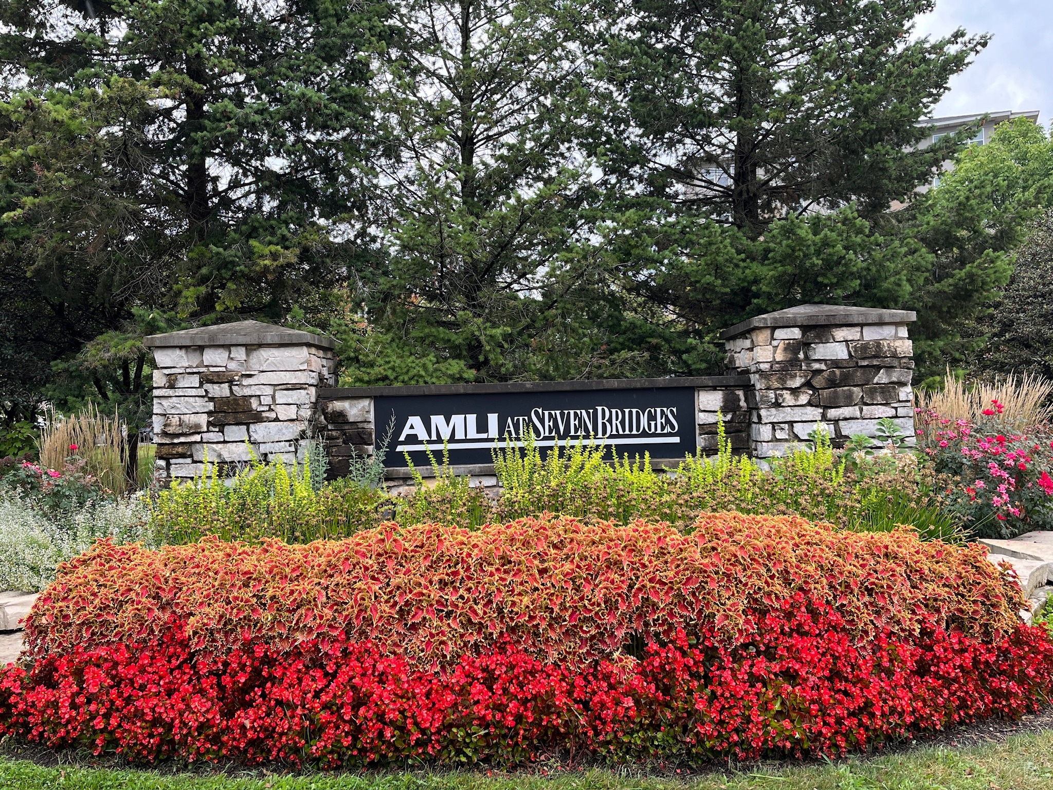 Stone entrance sign for "AMLI at Seven Bridges," surrounded by vibrant red flowers and lush greenery, set against tall evergreen trees.