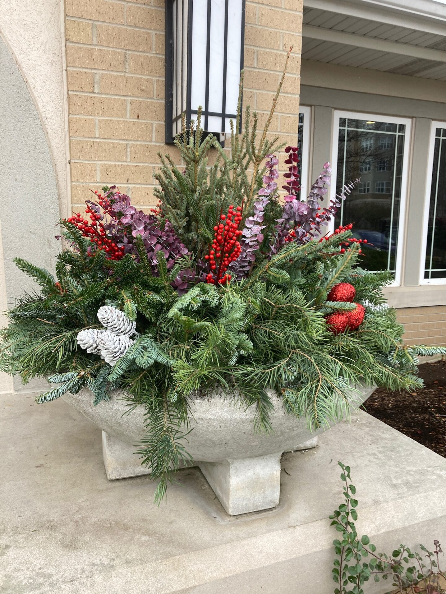 A large decorative planter with evergreen branches, red berries, and pinecones sits on a stone ledge outside a building with brick walls.