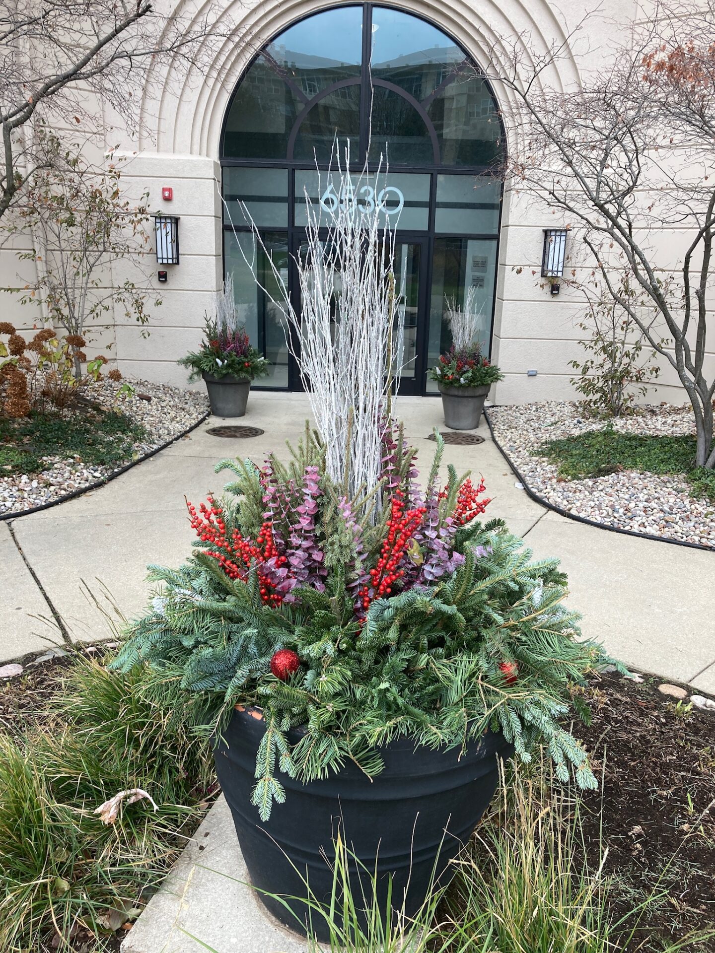 An arched entrance at 6830 features winter-themed decor with red berries and evergreen branches in a black planter, flanked by lanterns.