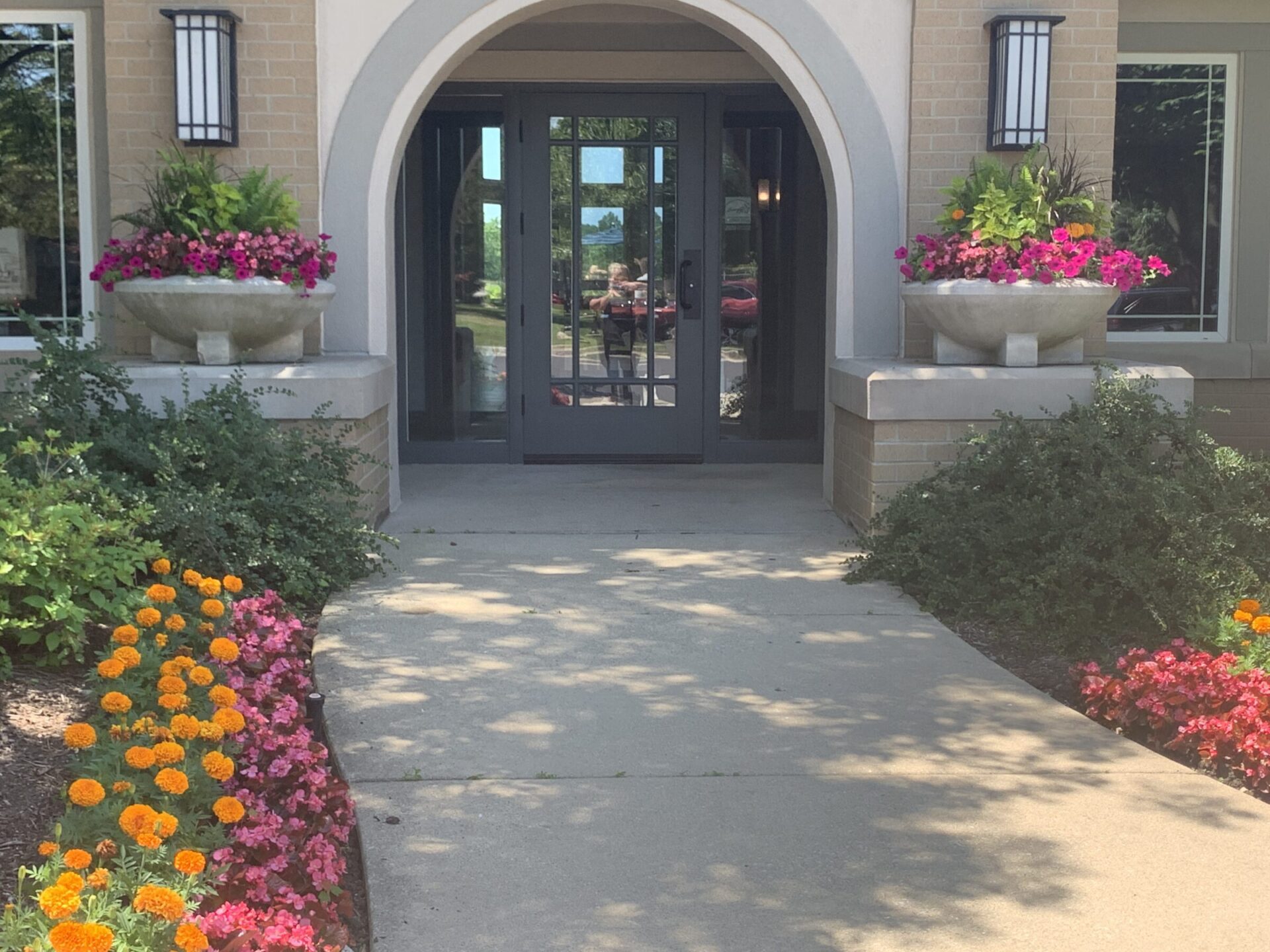 A landscaped building entrance with colorful flowers, decorative planters, and double doors. A person is reflected in the glass.