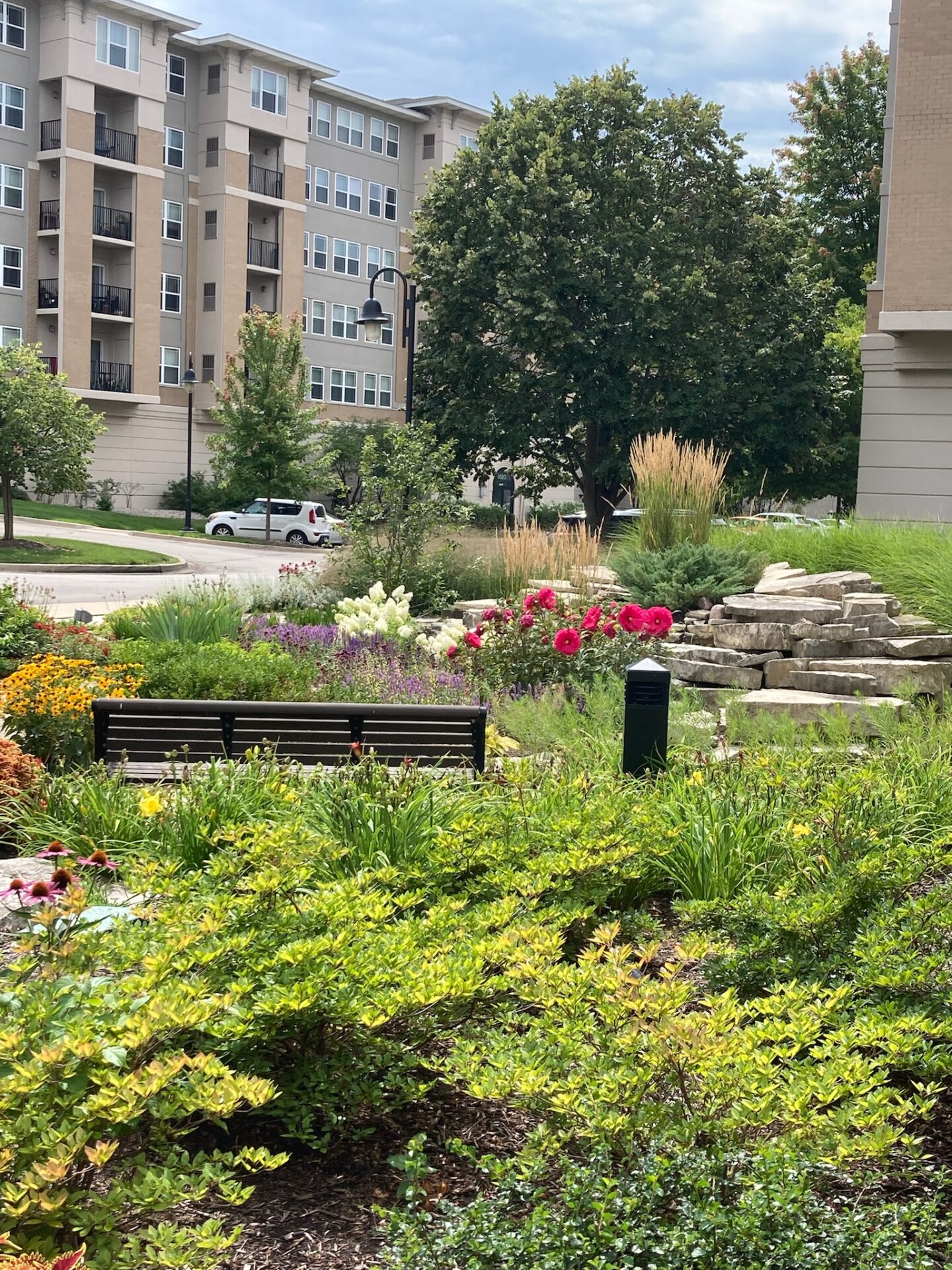 A serene garden with flowers and shrubs, adjacent to a modern apartment building. A bench overlooks the lush landscape and pathway.