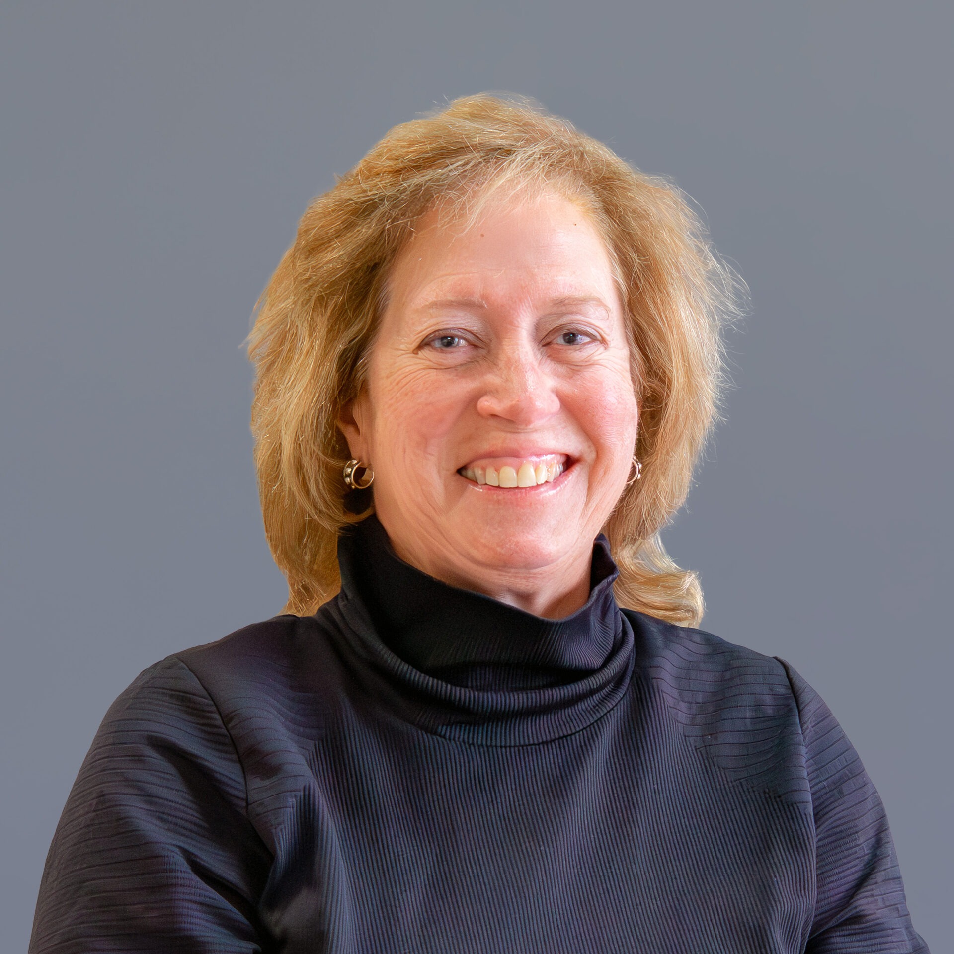 A smiling person wearing a dark top is captured against a plain gray background. The image focuses on the person's expression and attire.