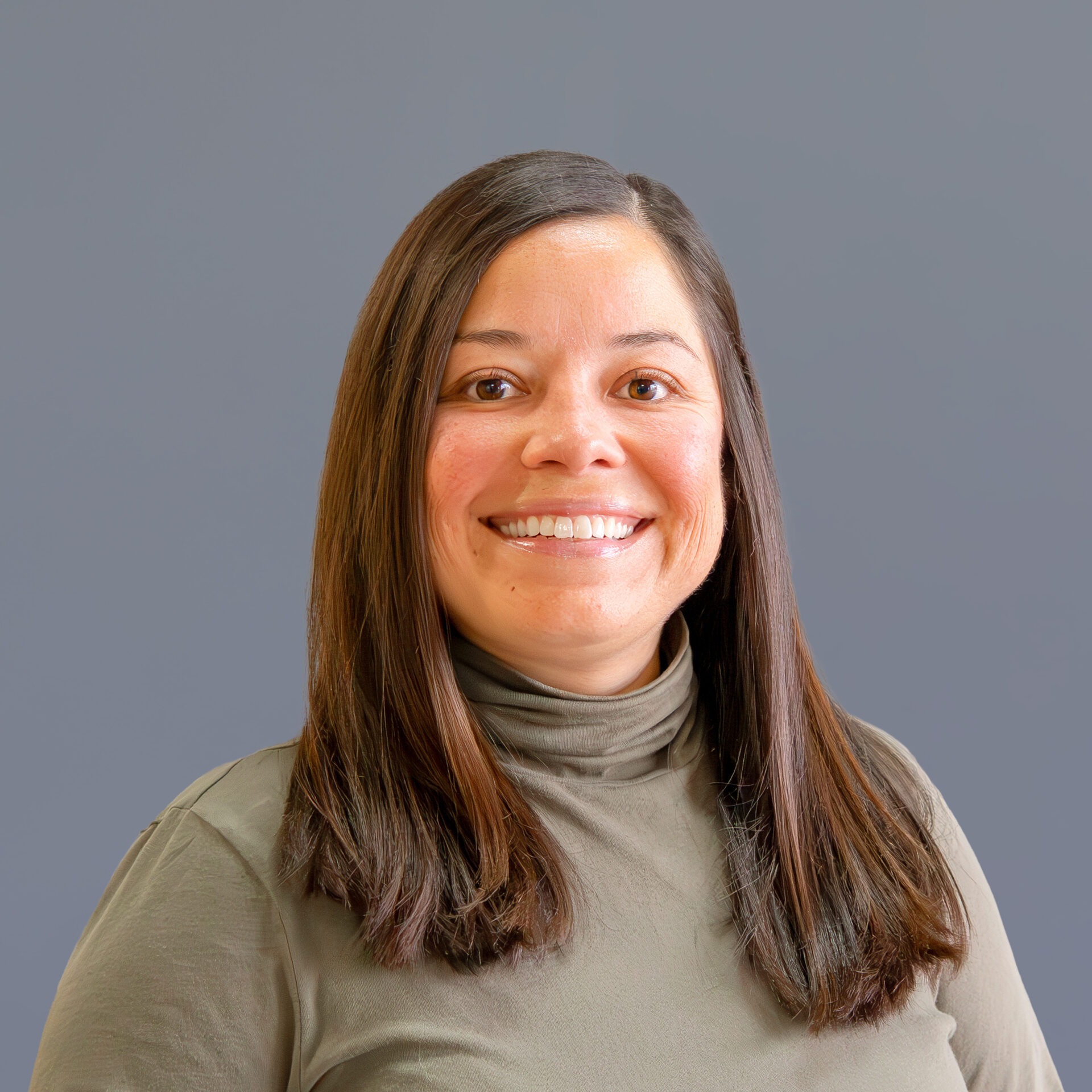 A person with long hair smiles against a plain gray background, wearing a green turtleneck. No landmarks or historical buildings are visible.