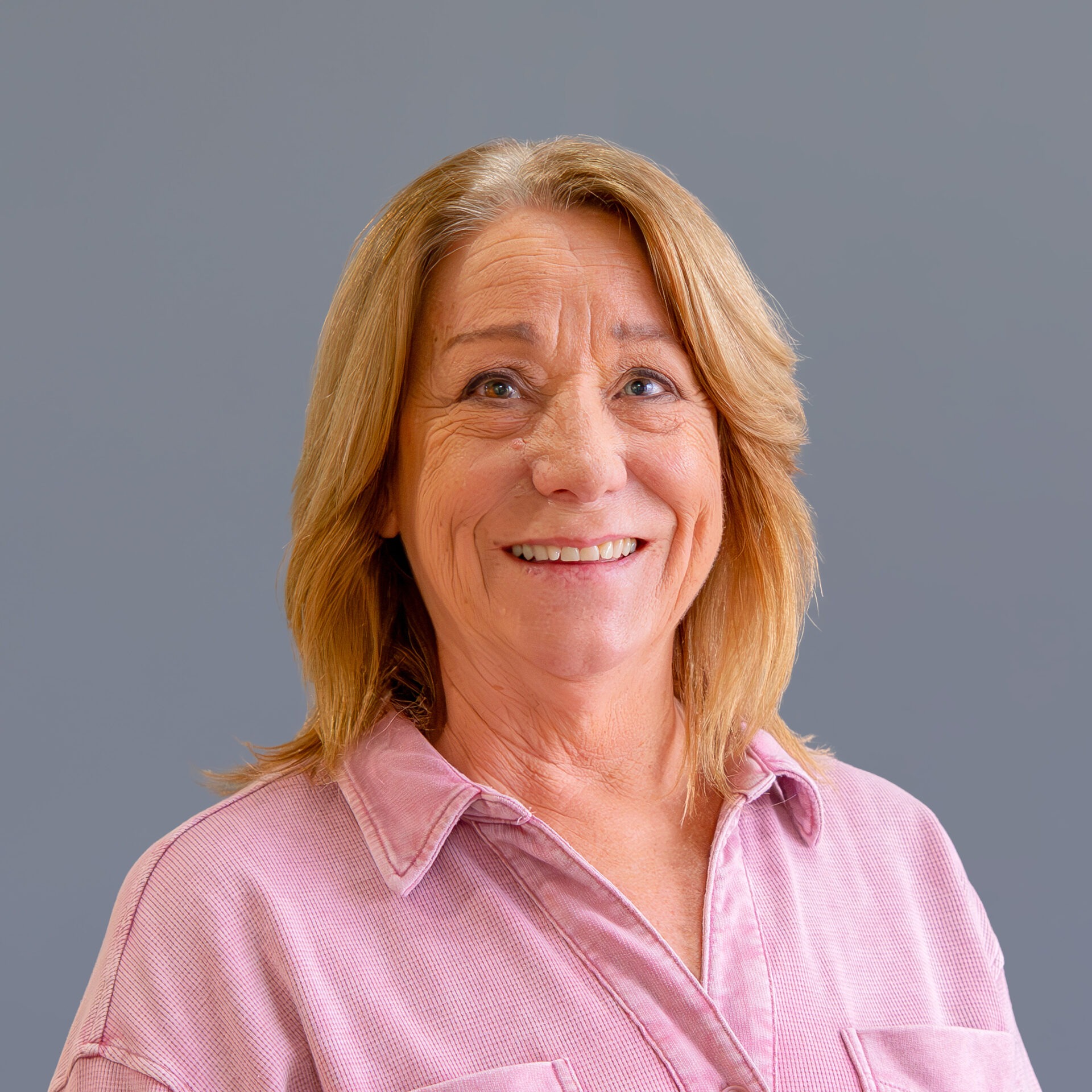 A smiling person with shoulder-length hair wearing a pink shirt stands against a plain gray background, creating a professional and approachable portrait.
