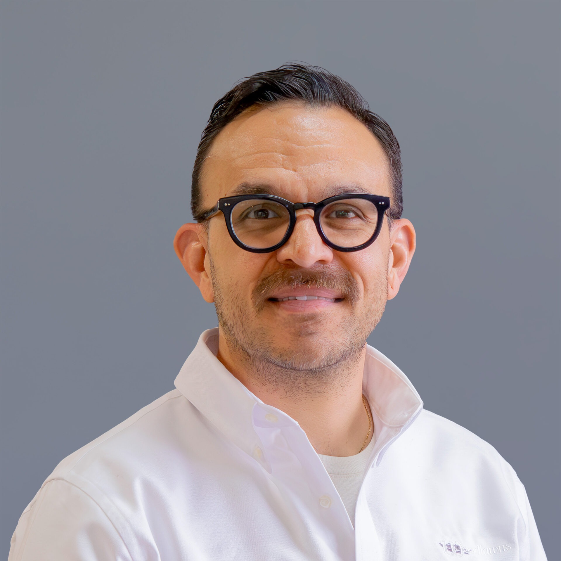 A person wearing glasses and a white shirt smiles against a neutral gray background. No landmarks or historical buildings are visible.