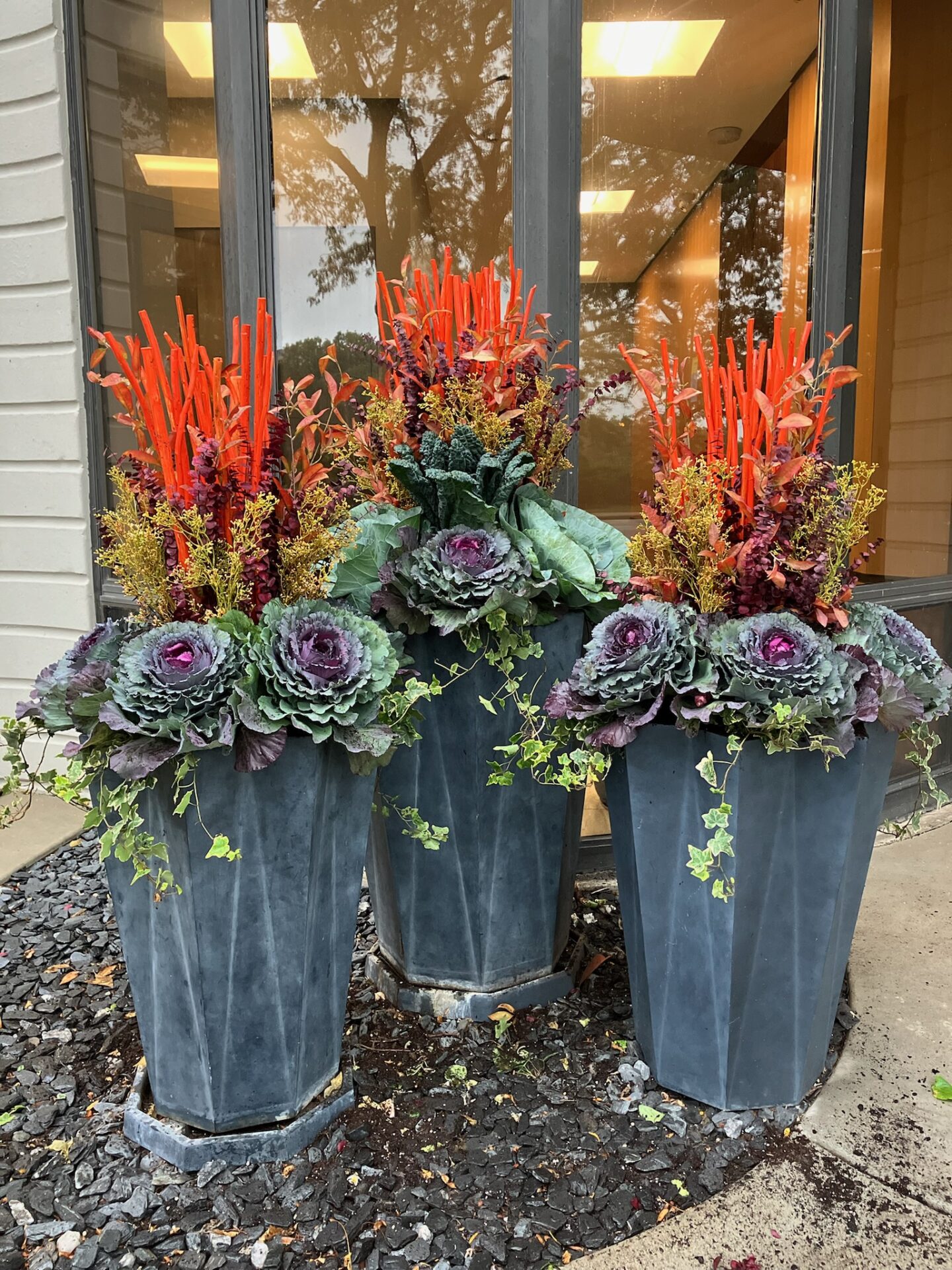 Three large planters hold vibrant arrangements of kale, red sticks, and yellow flowers, placed outside a building with reflective glass windows.