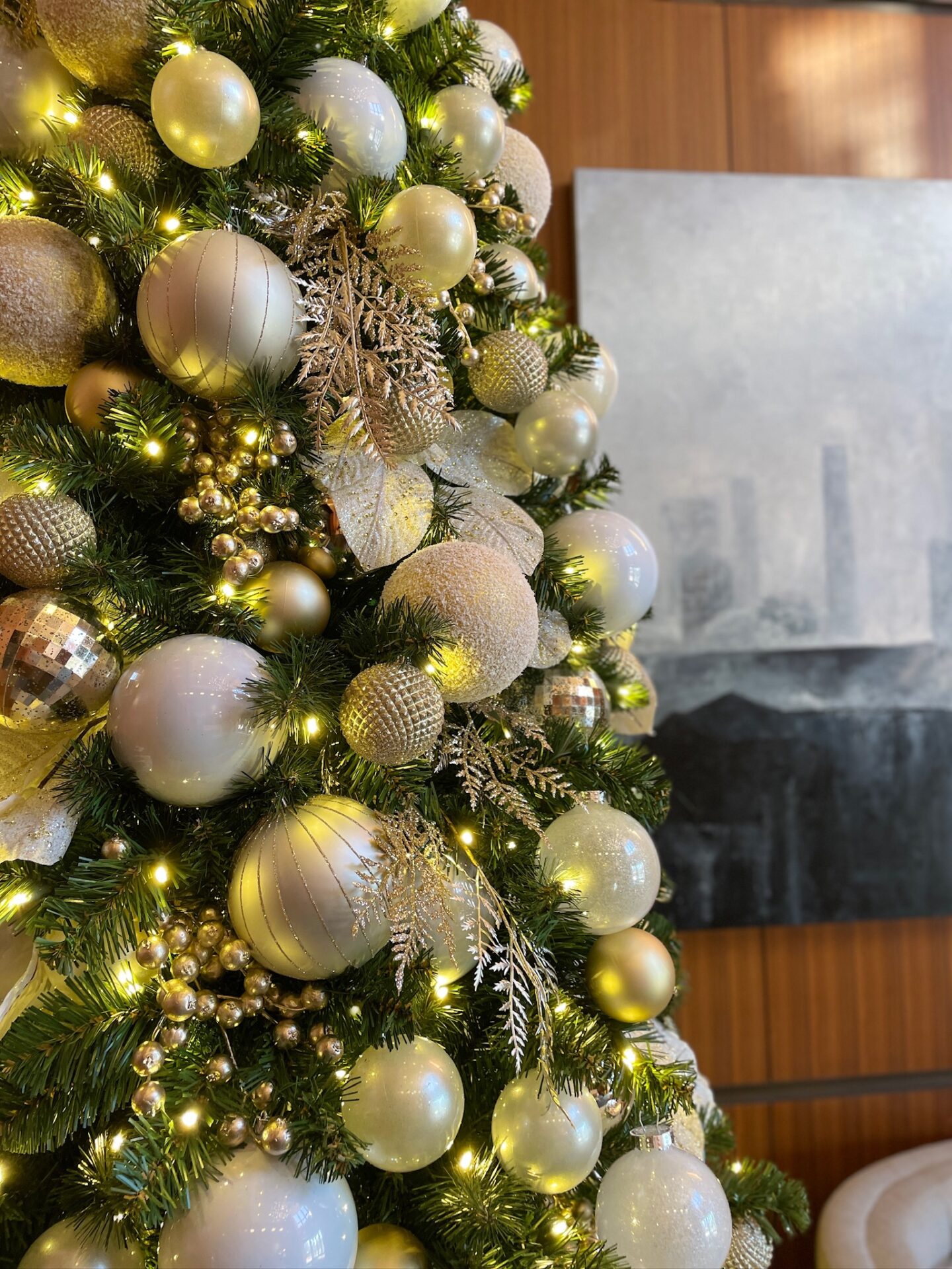 A beautifully decorated Christmas tree with gold and white ornaments, illuminated by soft lights. A blurred abstract painting is visible in the background.