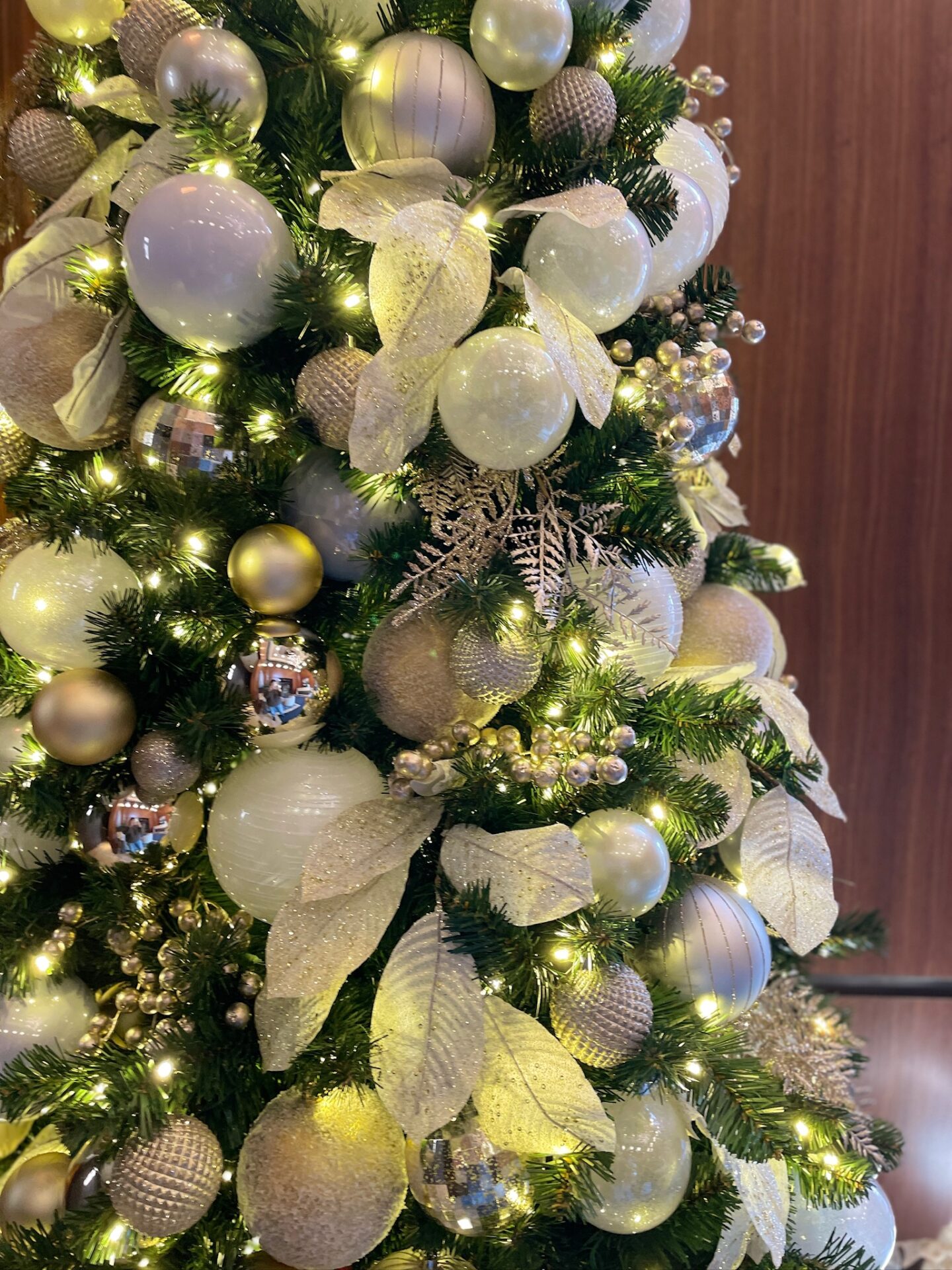 A close-up of a decorated Christmas tree with white and gold ornaments, ribbons, and sparkling lights against a wooden wall background.