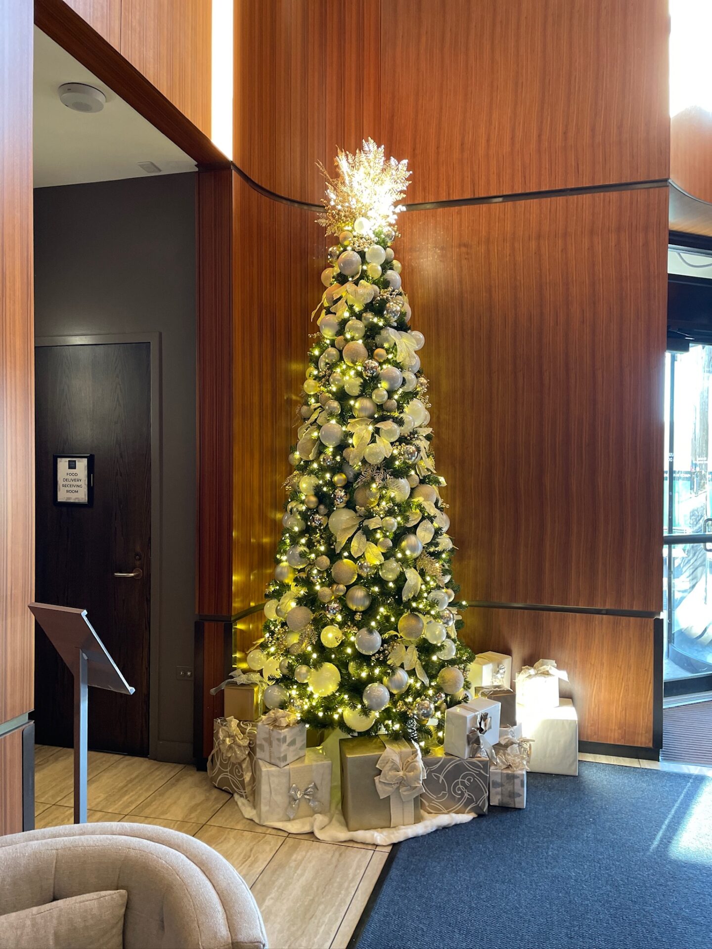 A beautifully decorated Christmas tree with white ornaments and lights, surrounded by wrapped gifts, stands in a warmly colored interior space.