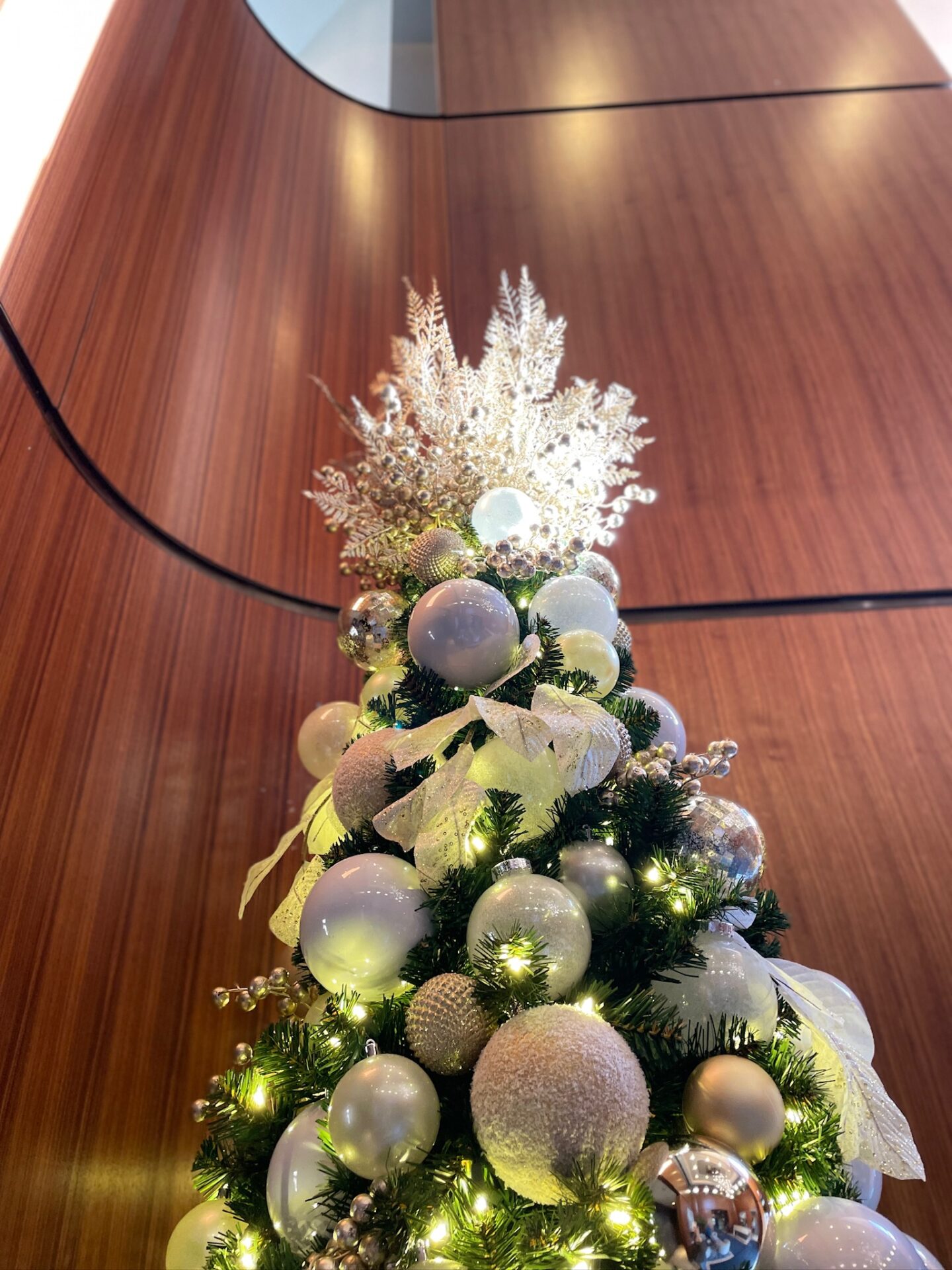 Elegant Christmas tree adorned with white and gold ornaments inside a modern wood-paneled room, creating a festive and sophisticated atmosphere.