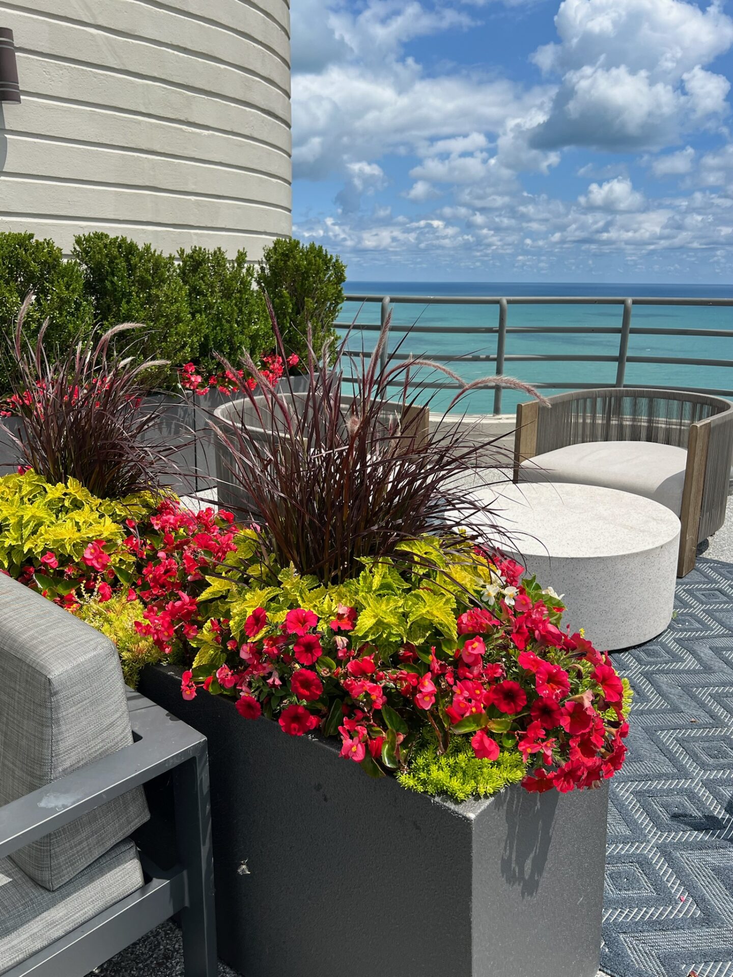Rooftop terrace with vibrant flowers, comfortable seating, and stunning ocean view beneath a partly cloudy sky. Peaceful and inviting ambiance.