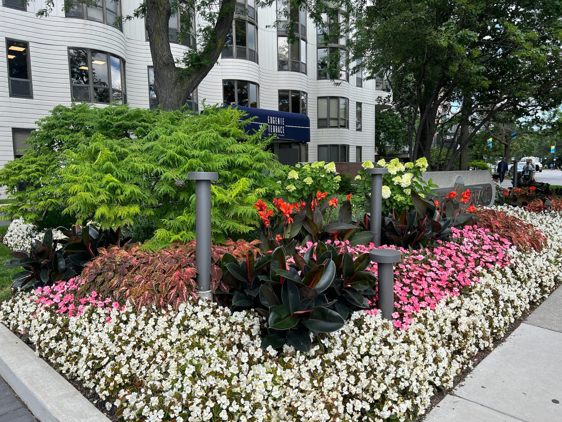 Lush garden with colorful flowers outside Eugene C. Eppley Terrace building. Two people walk on sidewalk, surrounded by trees and greenery.