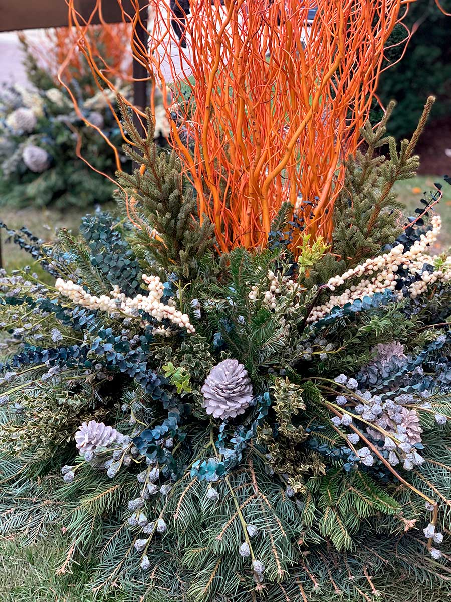 Vibrant autumn floral arrangement with orange branches, evergreen foliage, pine cones, and white berries, creating a striking natural display on a grassy area.