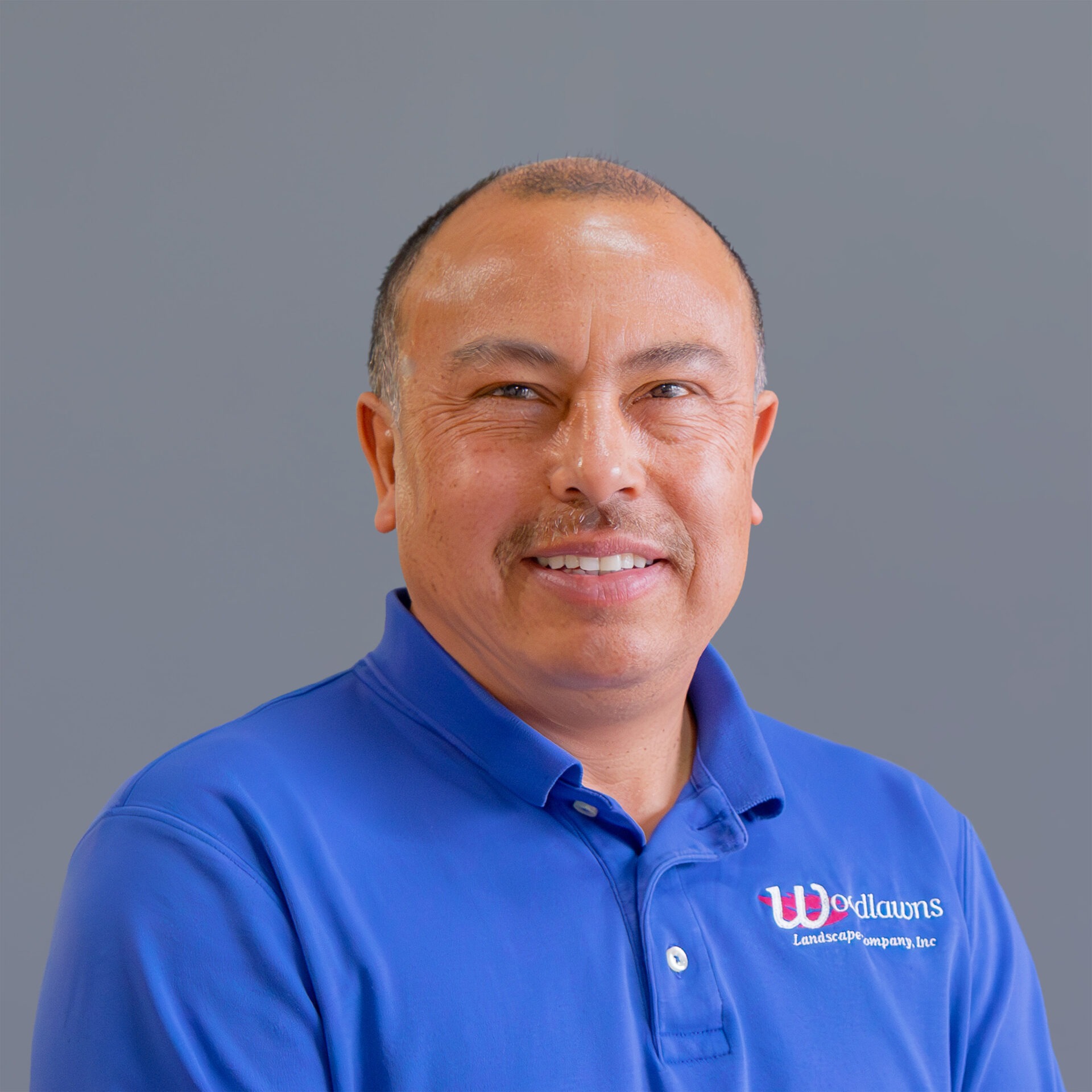 A person wearing a blue polo shirt with a company logo is smiling against a plain gray background.