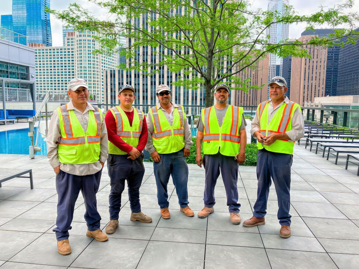Five persons in safety vests stand on a rooftop with a pool, surrounded by modern skyscrapers and buildings, under a clear sky.