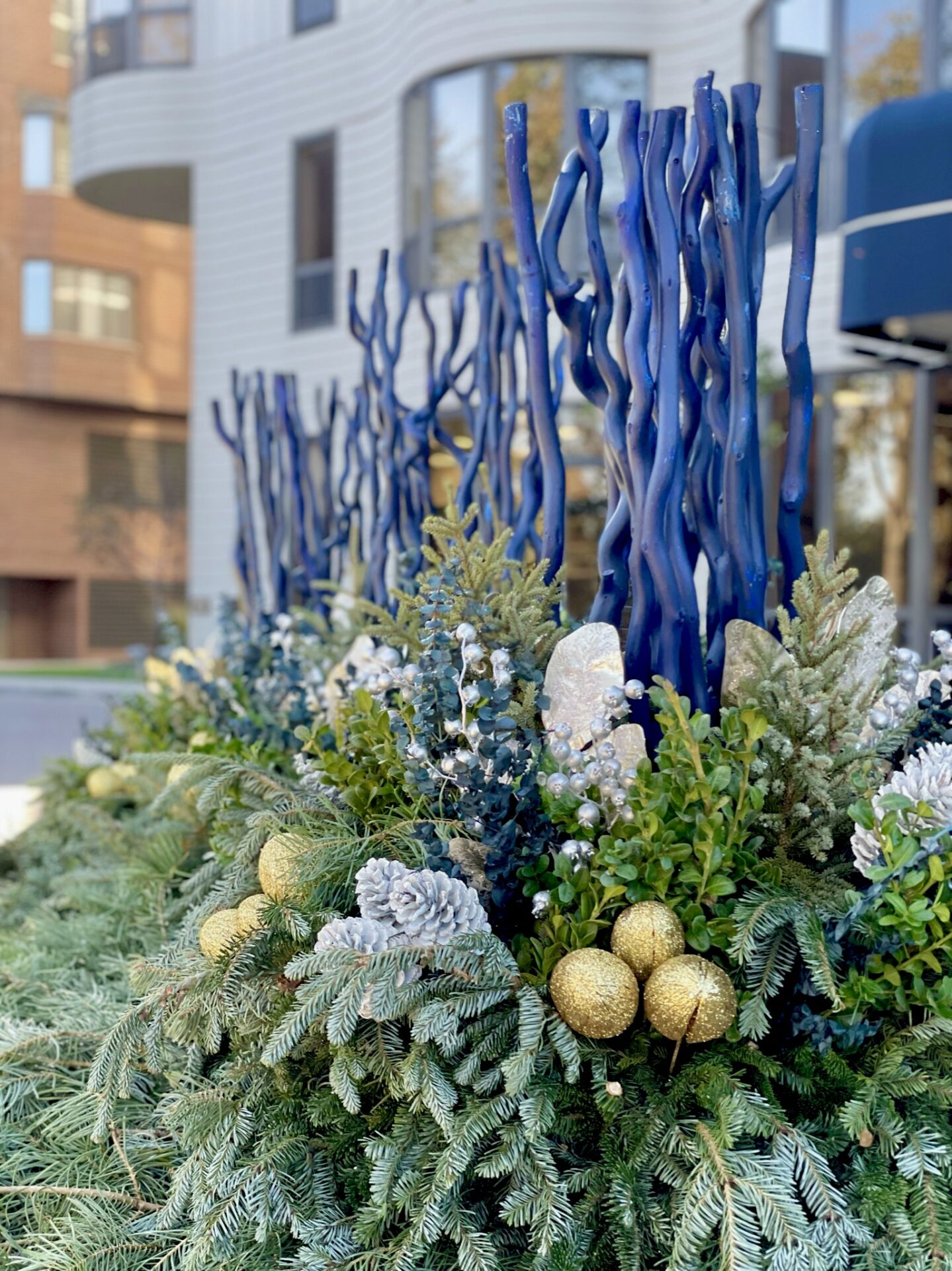 Blue decorative branches rise from lush greenery and gold ornaments, set against a modern building facade with curved windows in the background.