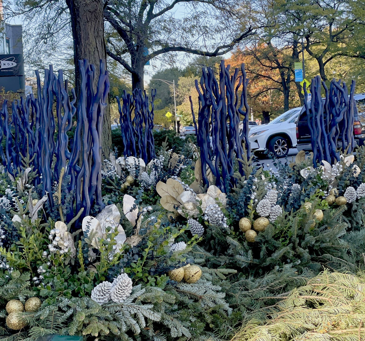 Decorative plants and blue sculptures adorn a city street with trees and vehicles. A person walks in the background, creating an urban scene.