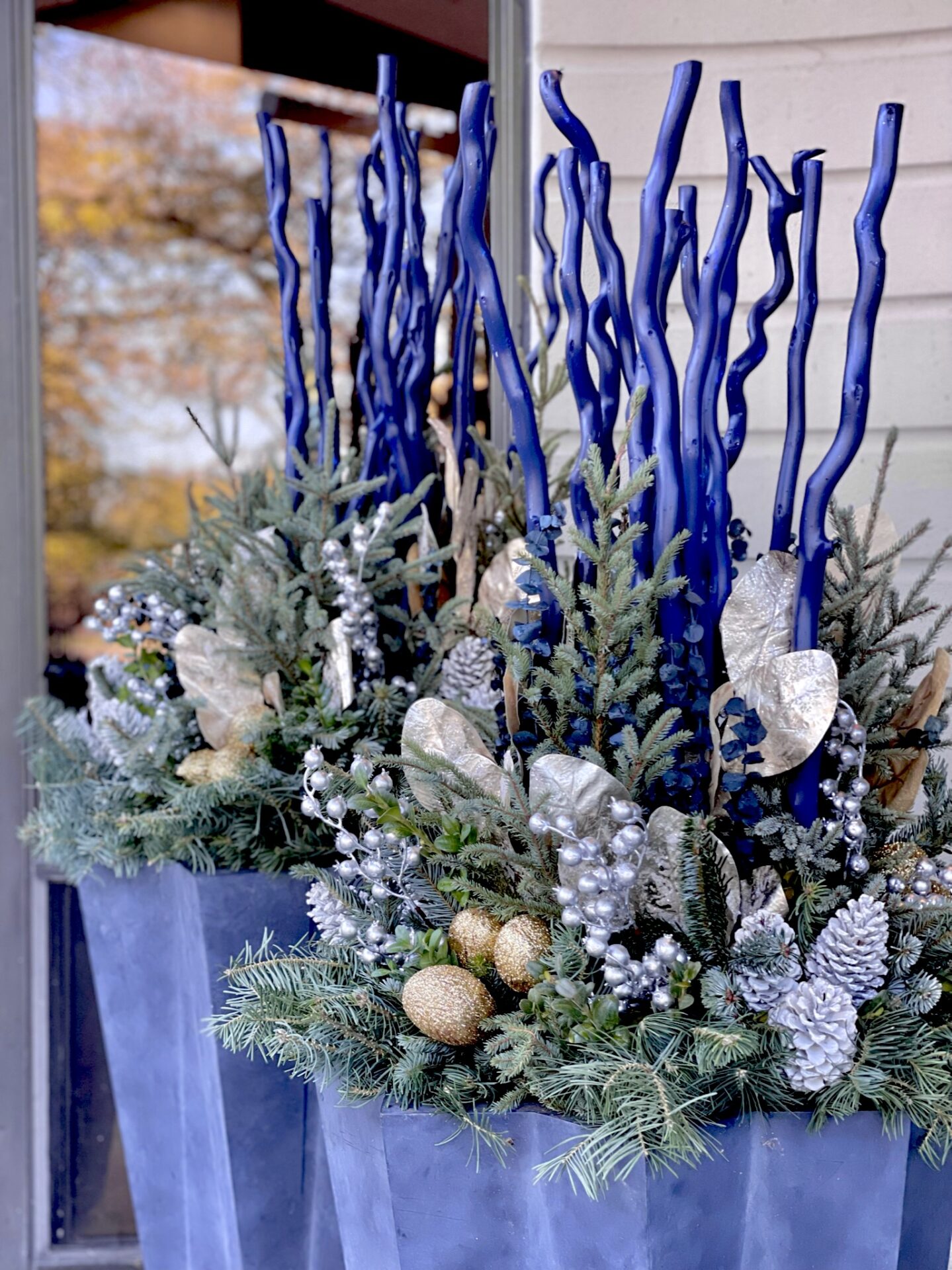 Festive blue and silver floral arrangement in large planters, adorned with pinecones and golden accents, set against a blurred outdoor backdrop.