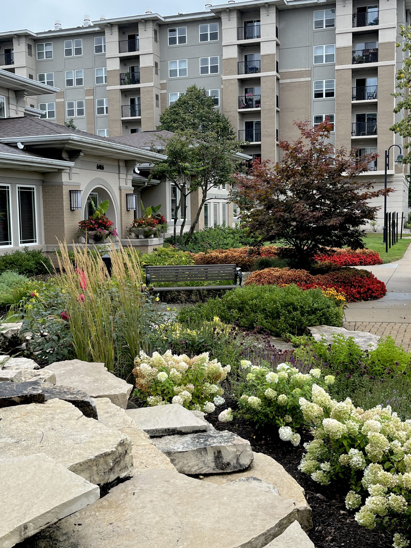 A landscaped garden with colorful flowers and bushes is set in front of a multi-story residential building on a cloudy day.