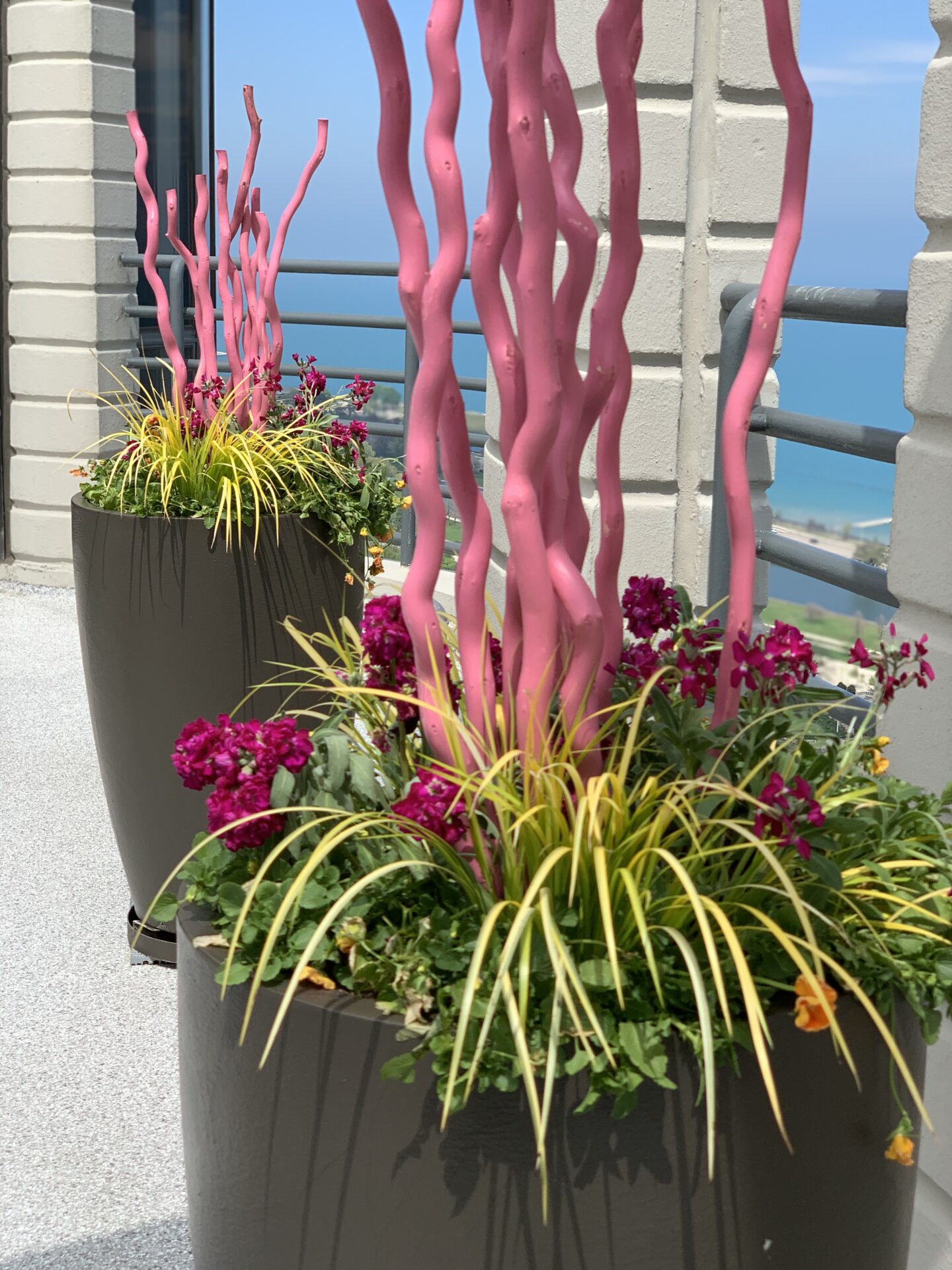 Colorful planters with pink installations and flowers decorate a balcony overlooking a blue ocean view under a clear sky.