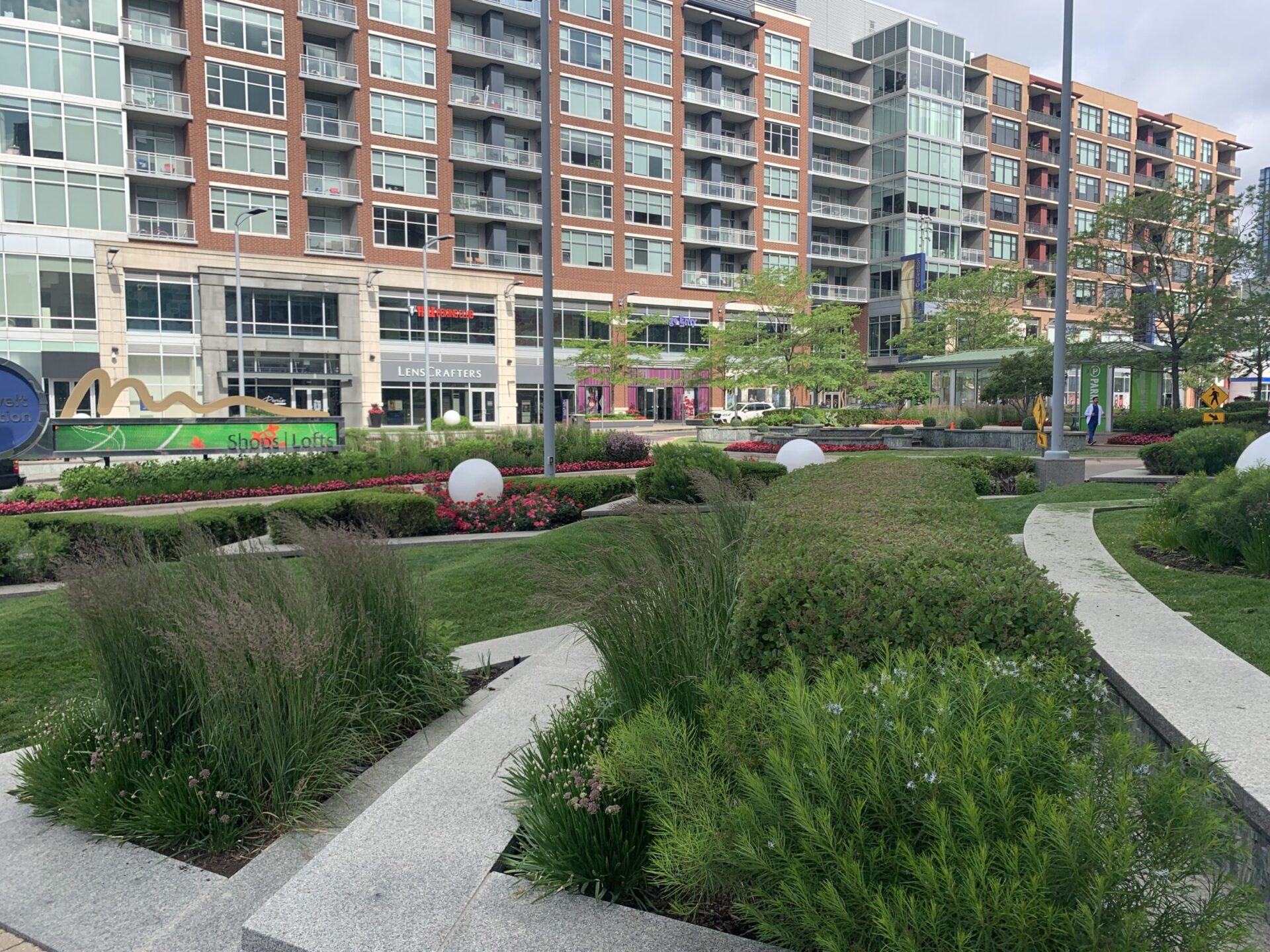 Urban park with greenery and pathways, bordered by modern apartment buildings. Signs for shops and a person walking are visible in the background.