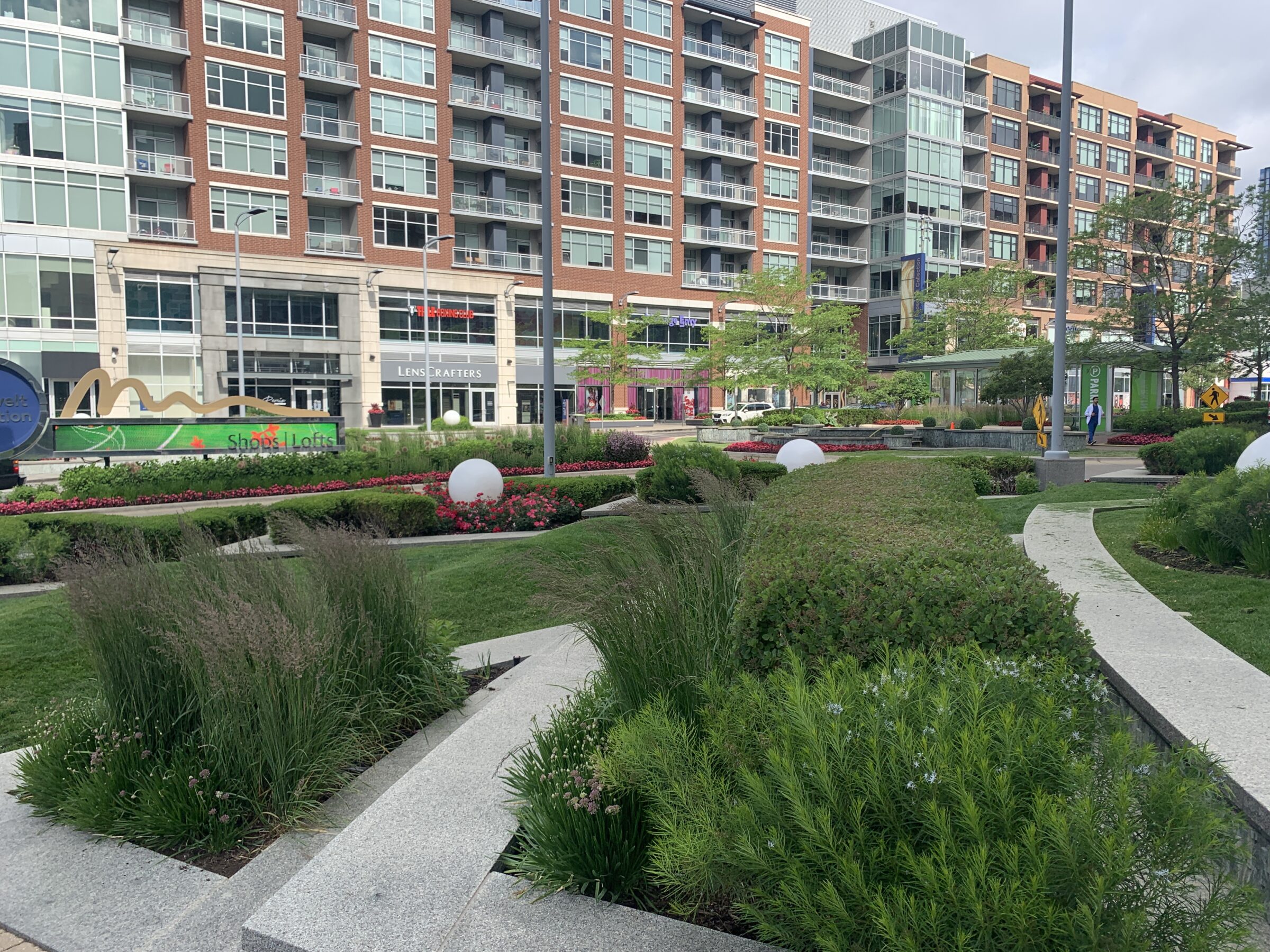 Urban park with greenery and pathways, bordered by modern apartment buildings. Signs for shops and a person walking are visible in the background.