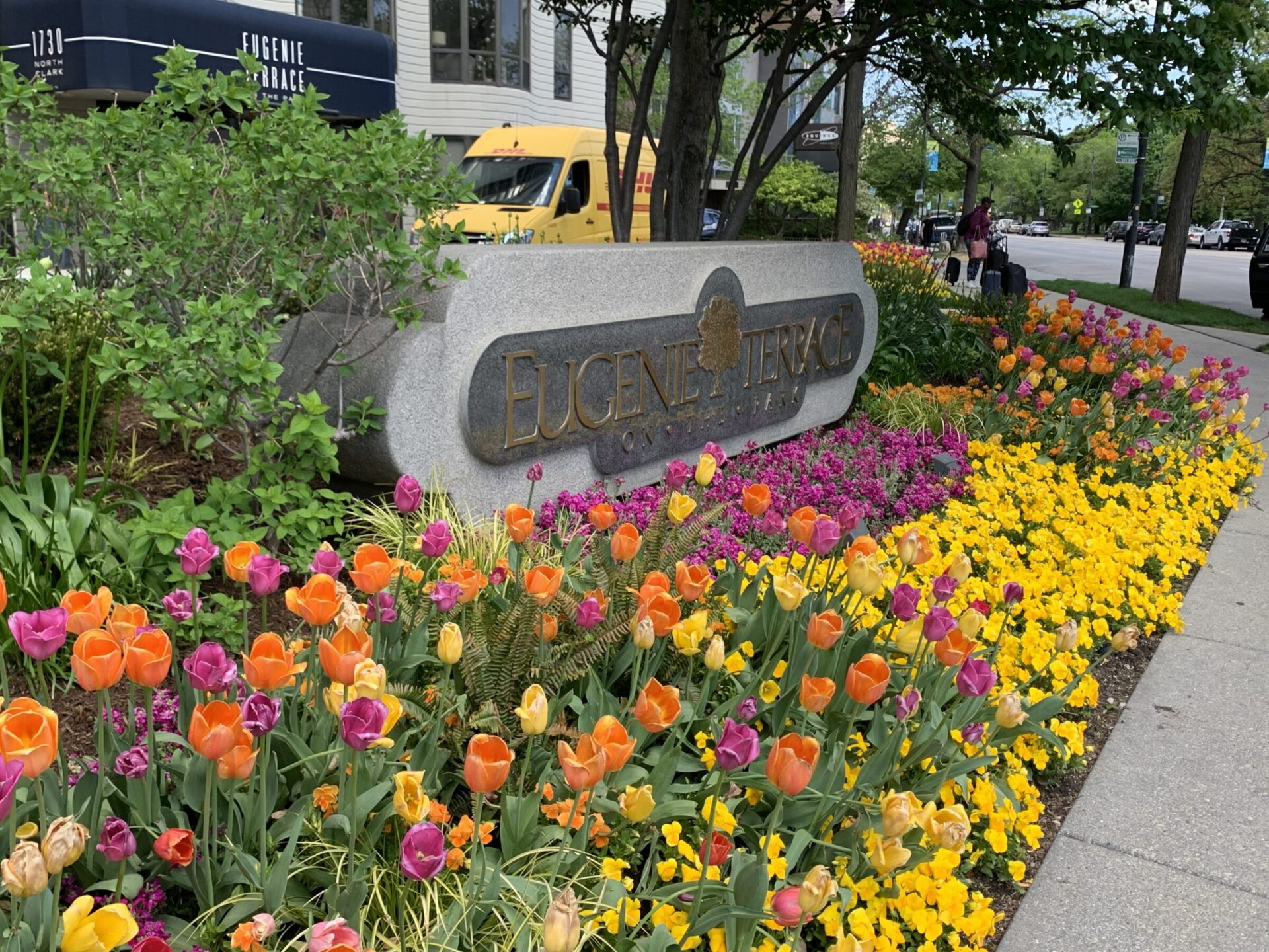 Colorful flowers surround the Eugenie Terrace sign near the sidewalk with a yellow vehicle and person in the background.