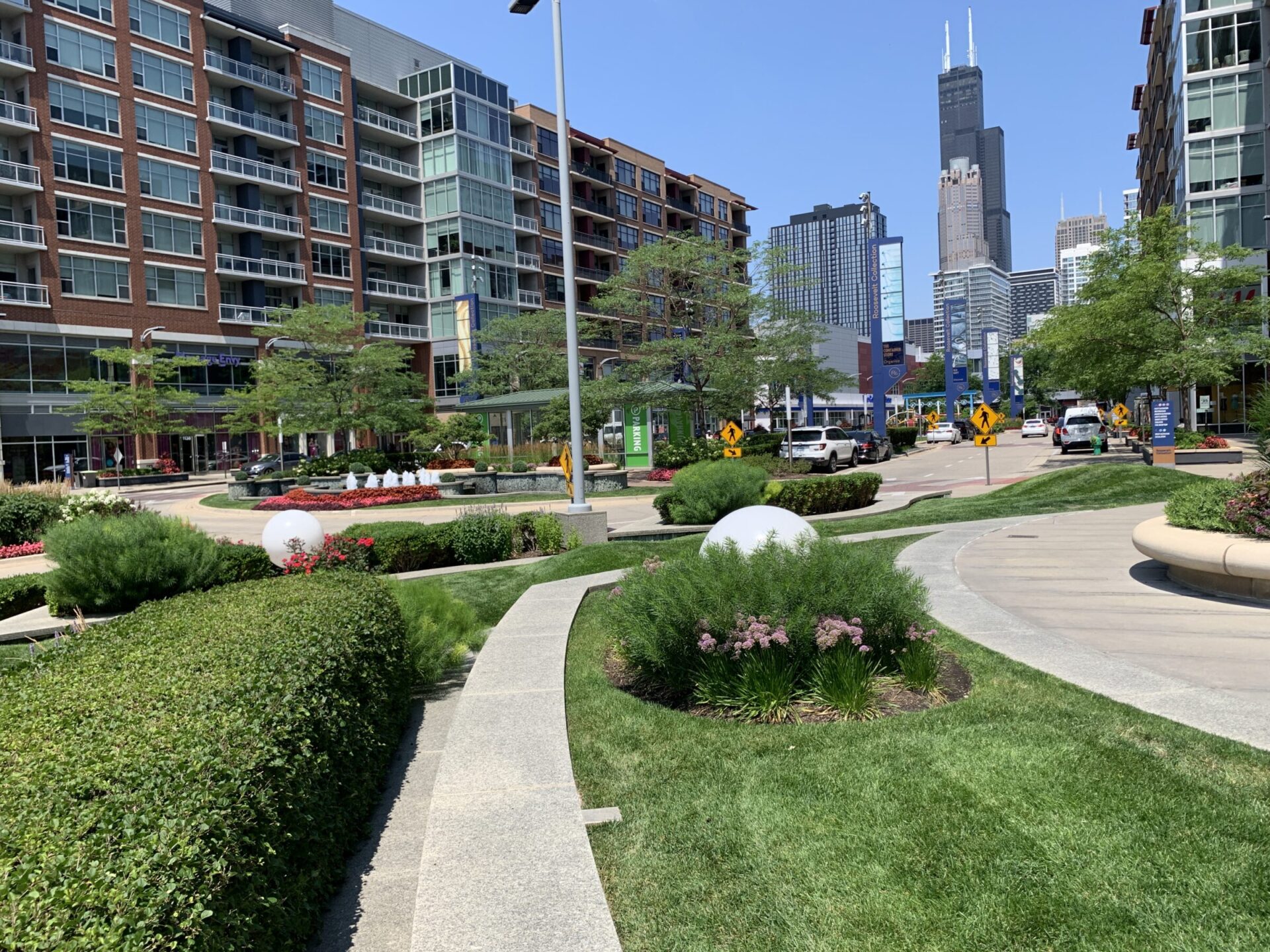 Urban park scene with modern buildings, cars, greenery, and people walking. Willis Tower visible in the background under clear blue sky.