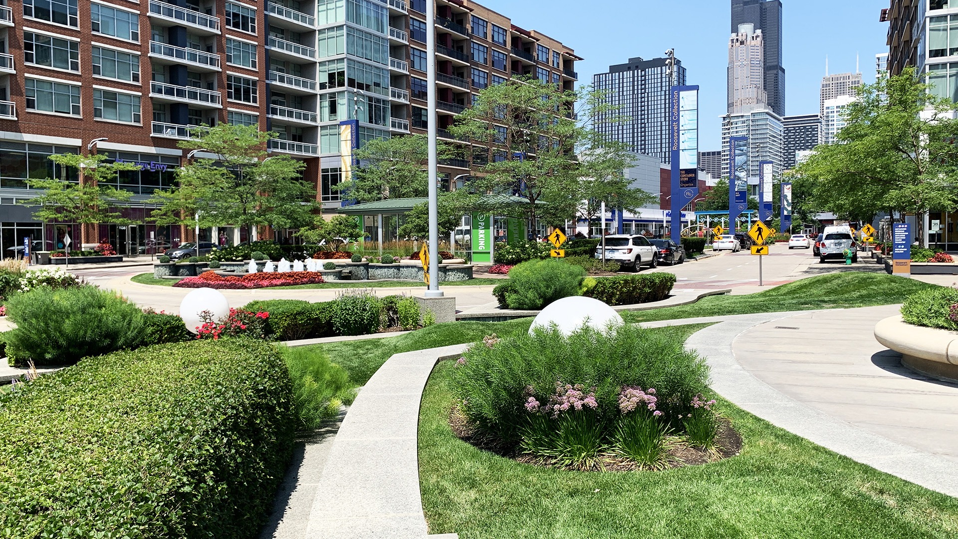 Urban park scene with landscaped greenery, modern buildings, and a clear view of the Willis Tower. People and cars in the background.