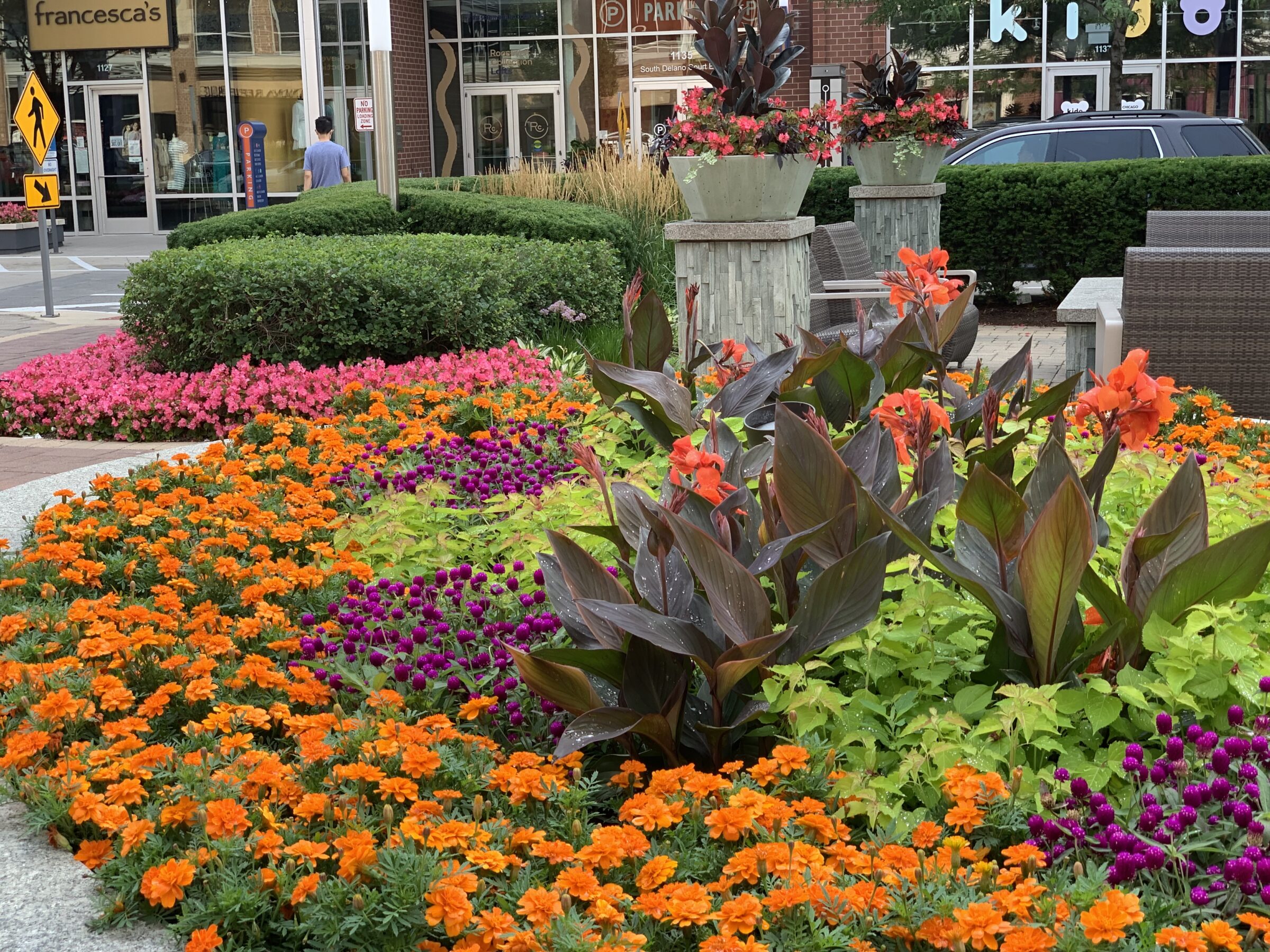 Colorful flowers and manicured bushes adorn a shopping area. A person walks by storefronts. Planters add to the vibrant, landscaped environment.