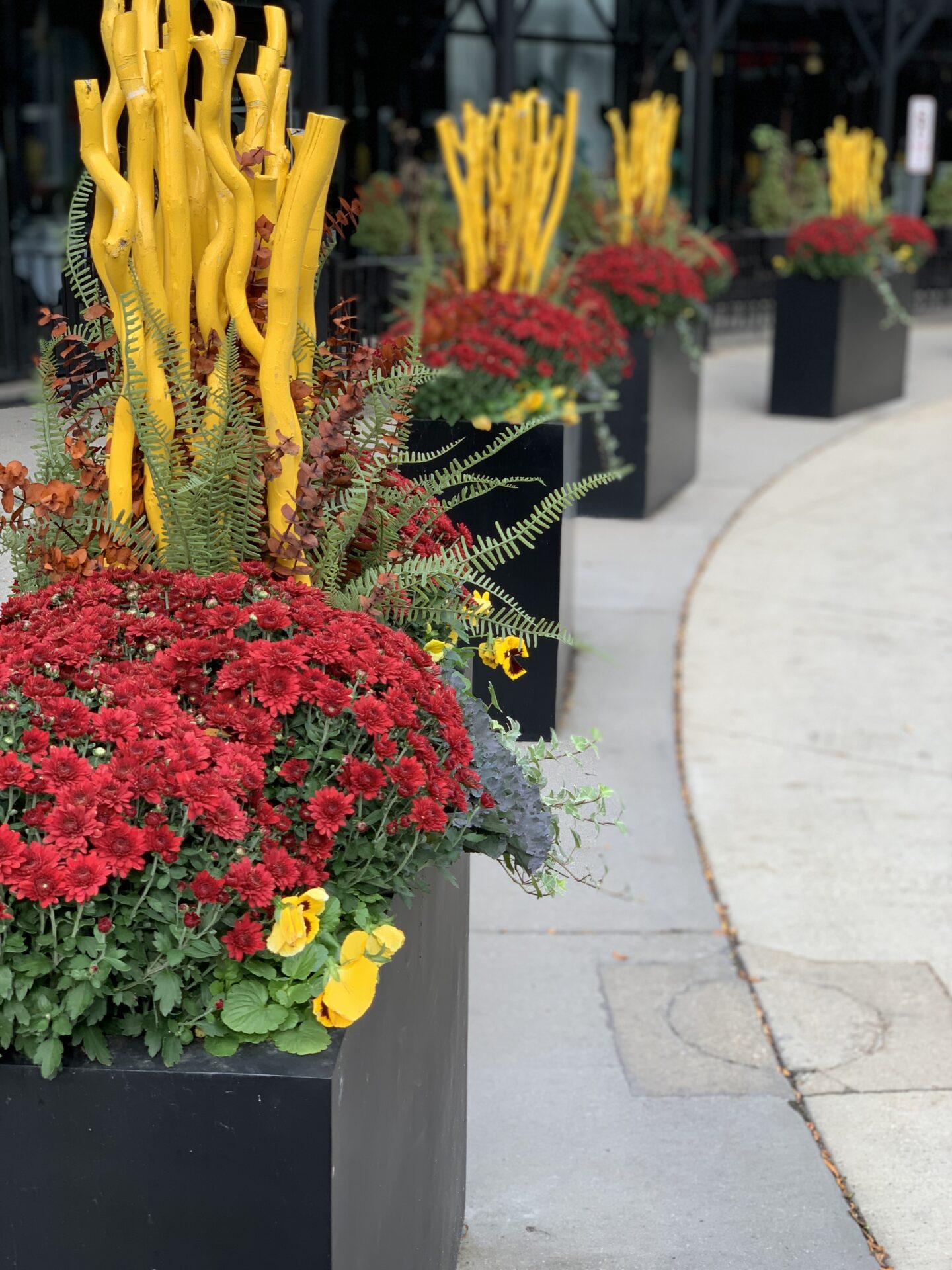 Vibrant street planters with red flowers and yellow sculptural sticks line a sidewalk near a building, adding a touch of whimsy.