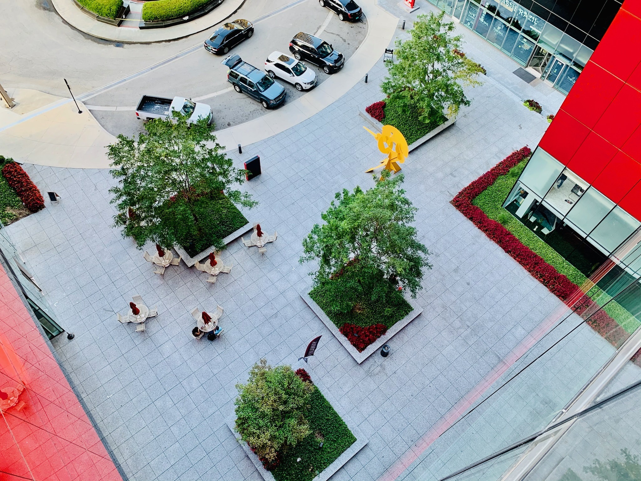 Aerial view of a courtyard with trees, seating areas, and a yellow sculpture, surrounded by red and glass buildings, and nearby parked cars.