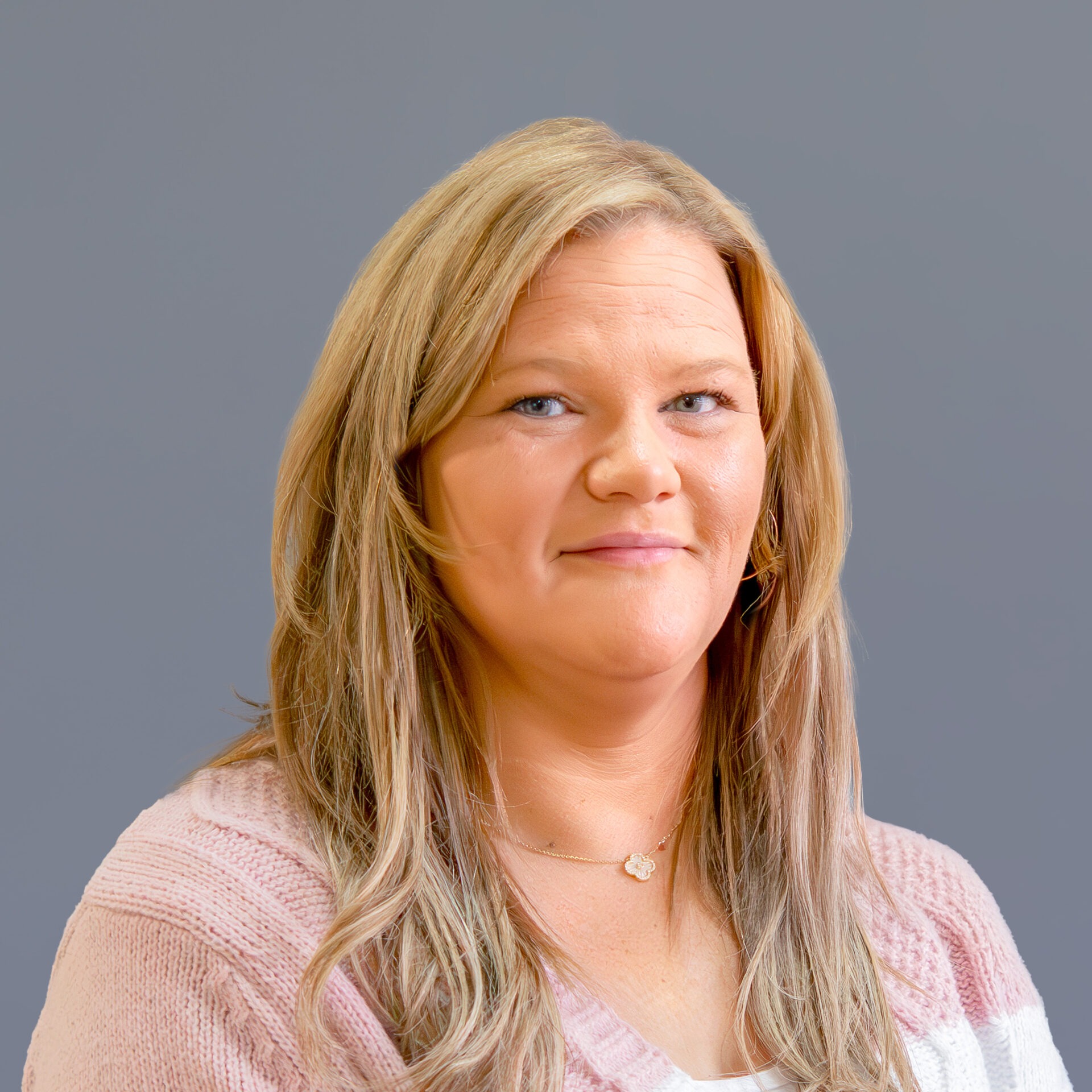 A person with long hair and a neutral expression is wearing a light pink sweater against a plain gray background.
