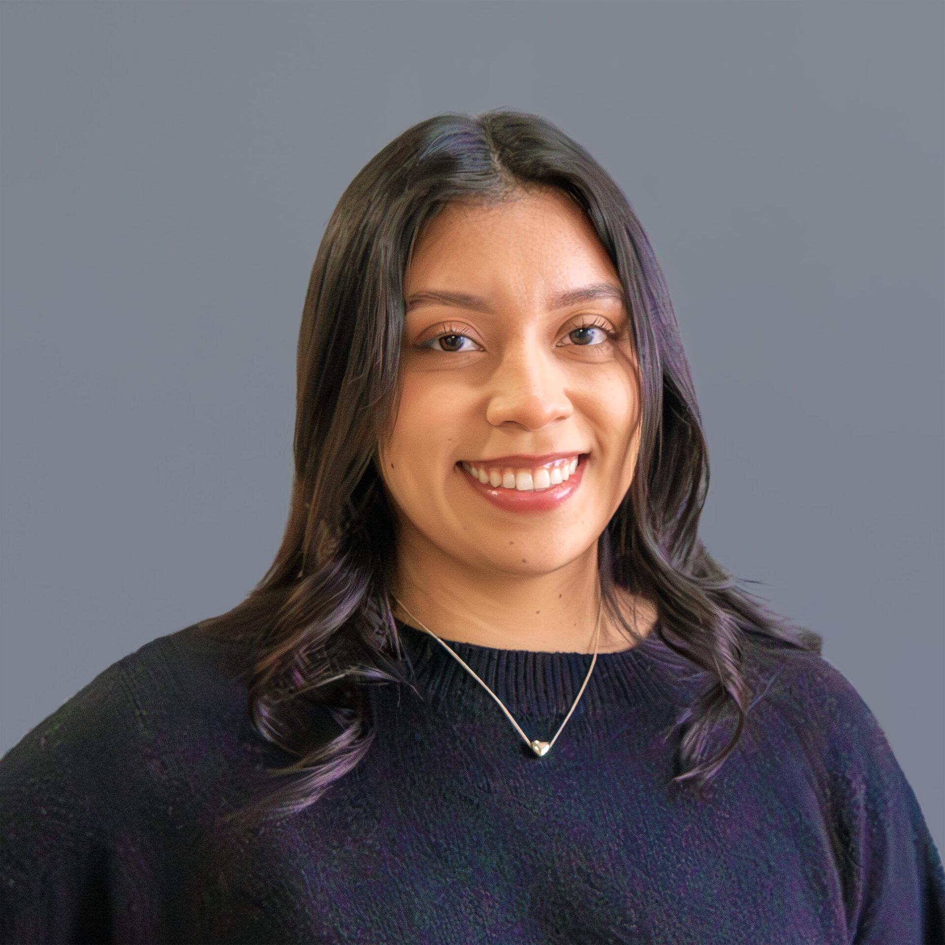 A person with long dark hair and a necklace smiles against a plain gray background, wearing a dark sweater.