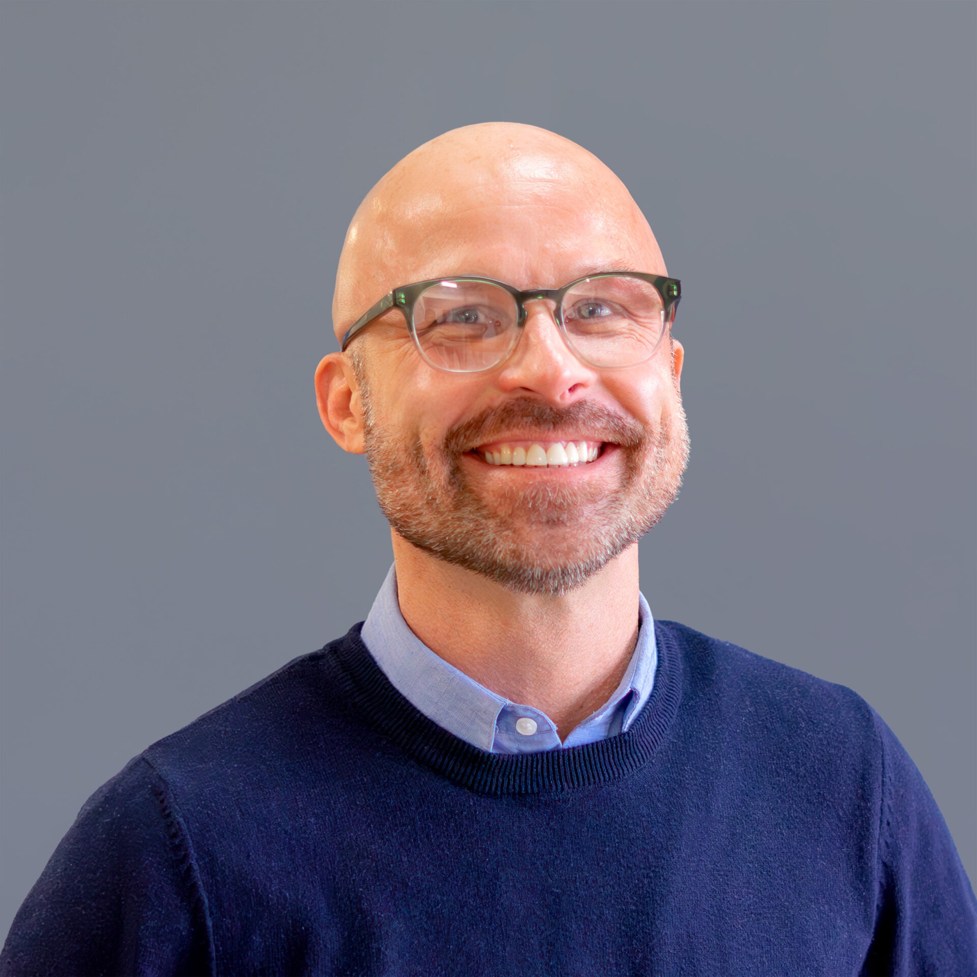 A smiling person with glasses and beard wears a blue sweater over a collared shirt, against a plain gray background.