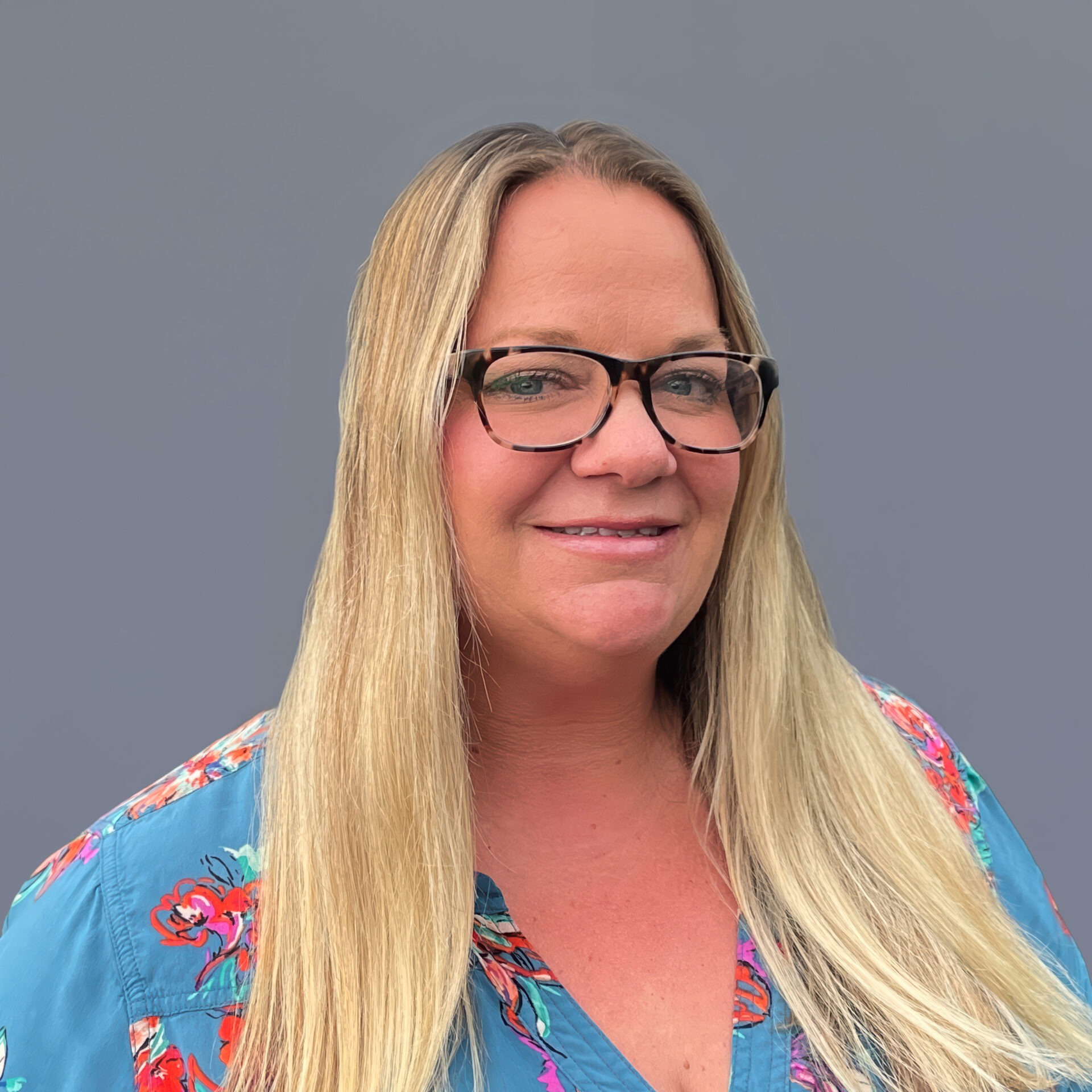 A person with long blonde hair and glasses, wearing a colorful floral shirt, stands against a plain gray background, smiling slightly.