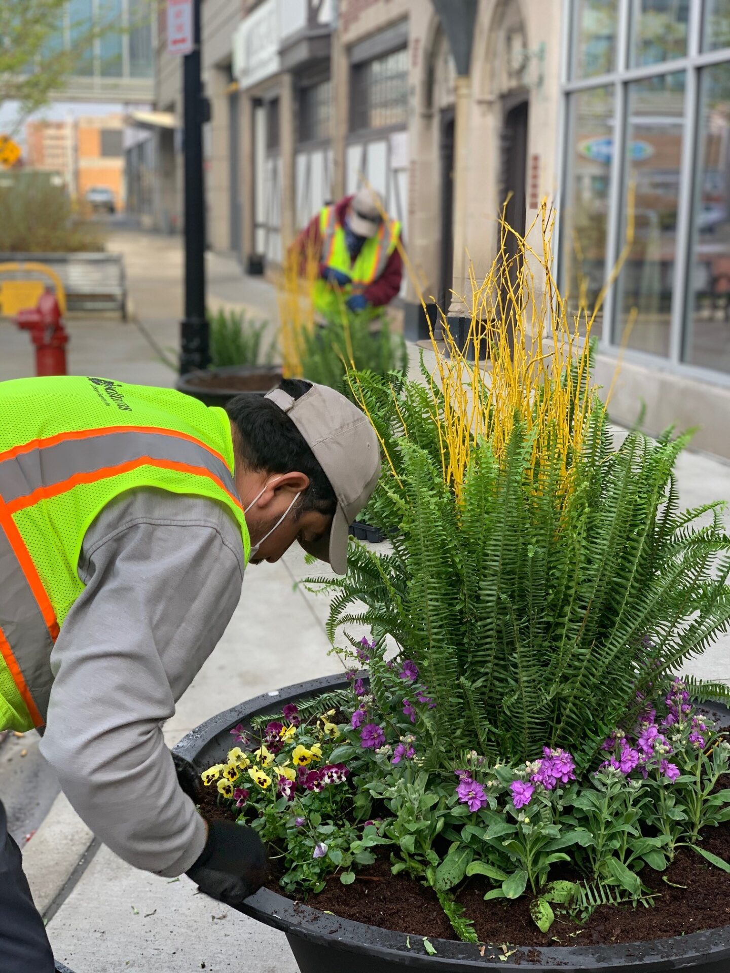 Two persons in safety vests plant flowers in large pots along a city sidewalk, near storefronts and brick buildings.