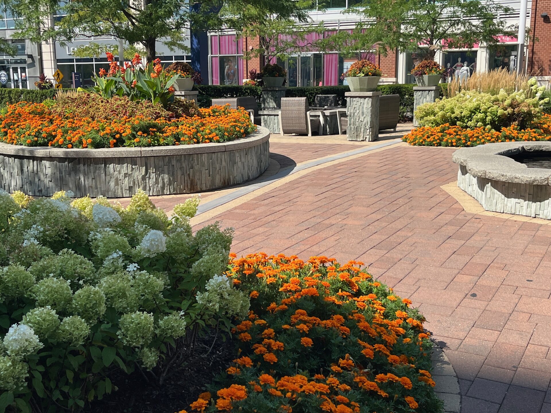 A vibrant, sunlit plaza features colorful flower beds, trees, modern planters, and a seating area with tables and chairs, surrounded by urban buildings.