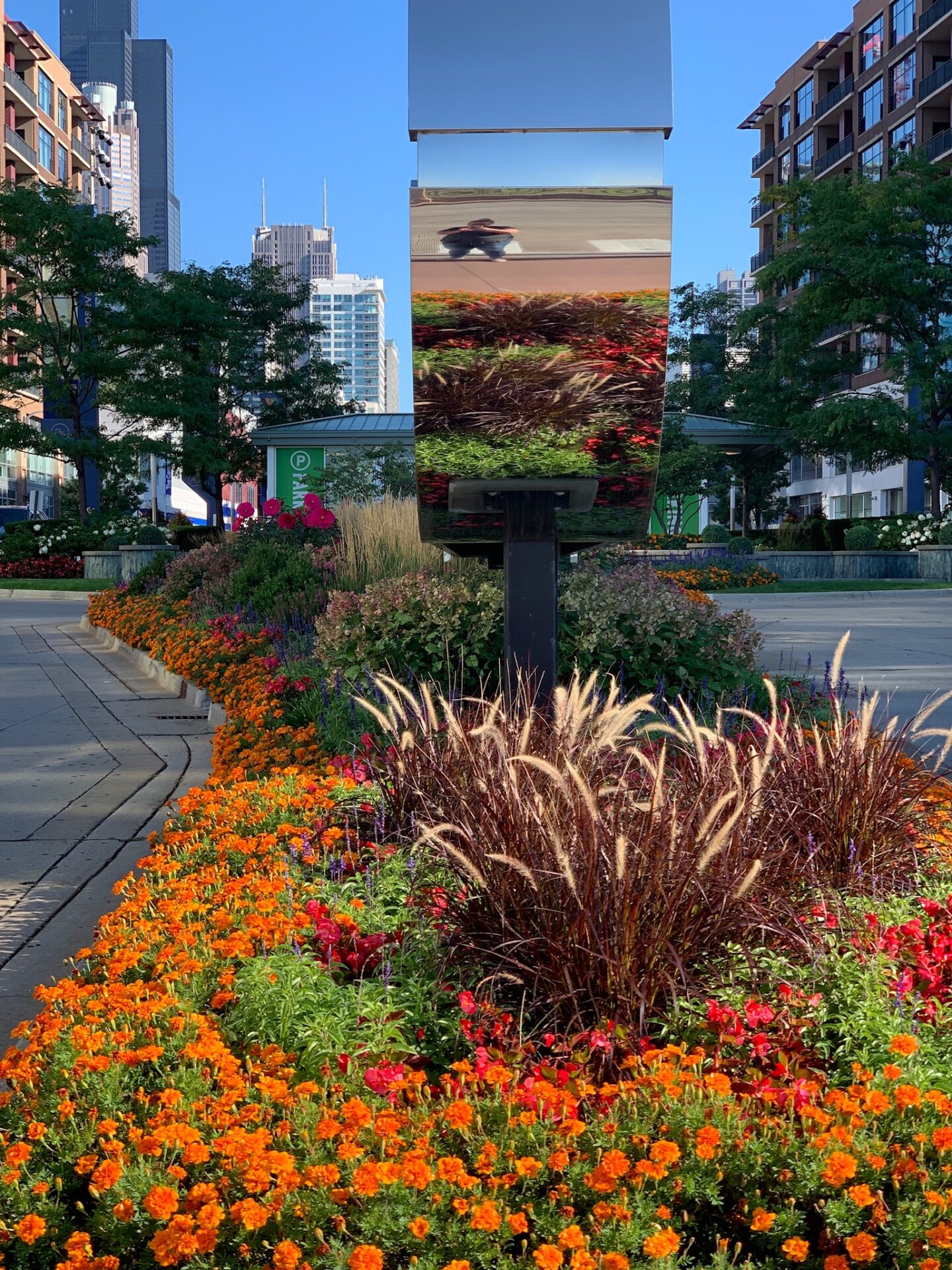 A vibrant garden surrounds a reflective structure, with Willis Tower in the background, amidst urban buildings and a pathway.