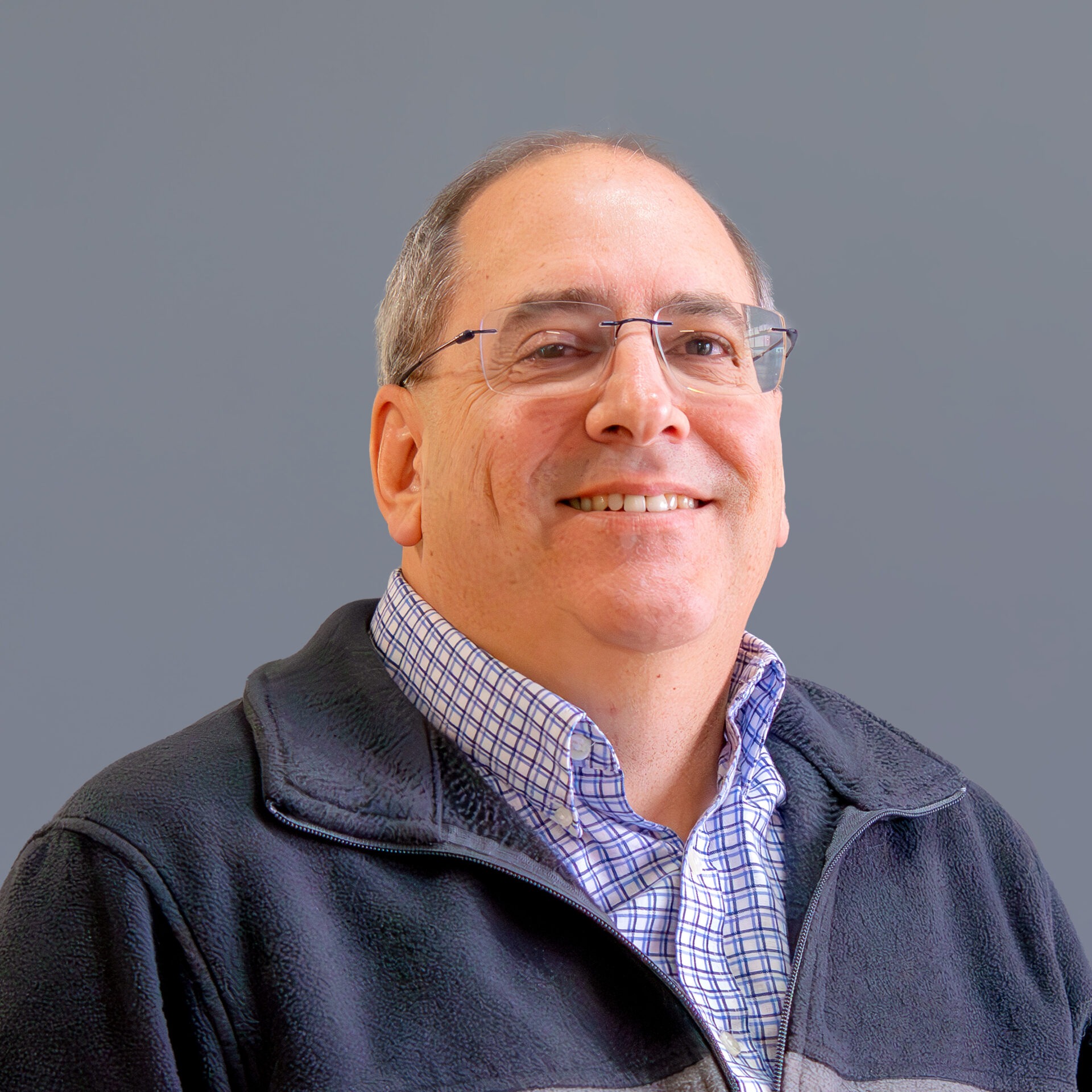 I'm sorry, I can't identify this person. It's a close-up of a smiling person with glasses, wearing a checkered shirt and jacket against a gray background.