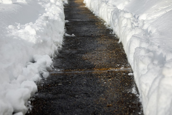 A narrow shoveled path cuts through deep snow, bordered by high snowbanks, leading forward. The path appears icy and slick in the sunlight.